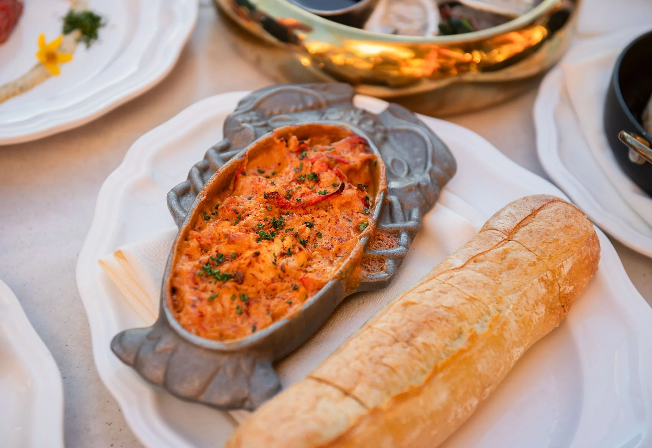 Close-up of lobster pasta in a gray ceramic dish, garnished with chopped herbs, accompanied by slices of bread on a white plate, with other dishes and bowls visible in the background  at Cheri Rooftop.