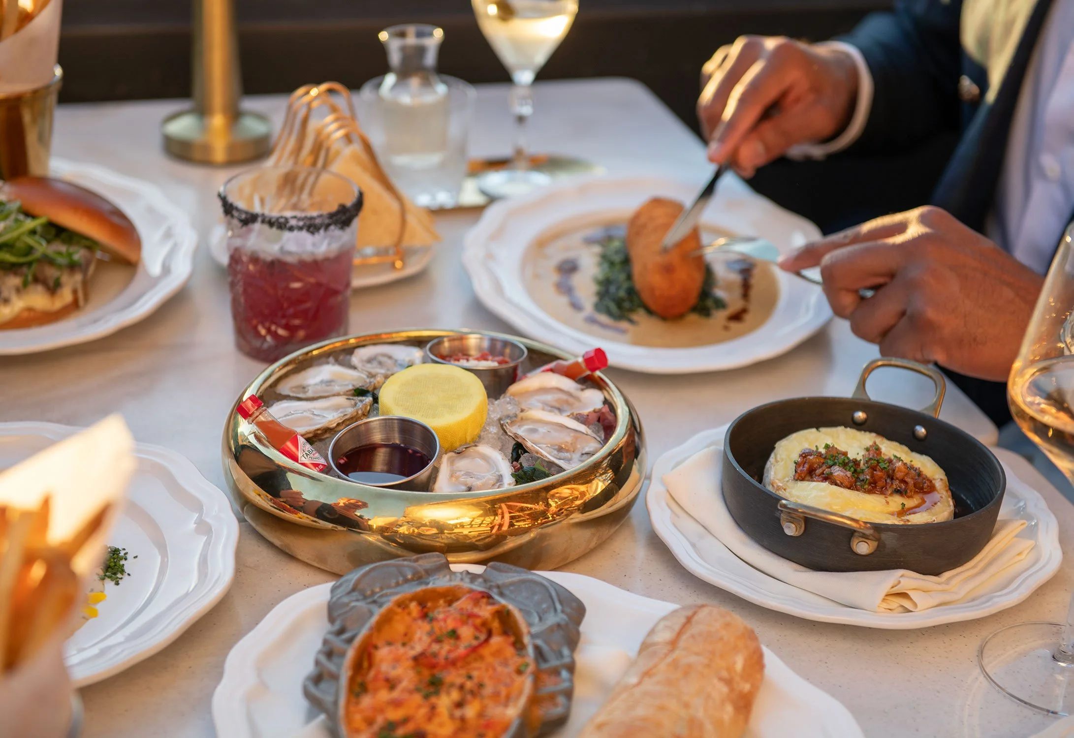 A table filled with various seafood dishes including oysters, a crab cake, and other plated entrees, along with wine glasses and a beverage with a salted rim, in a dining setting  at Cheri Rooftop.