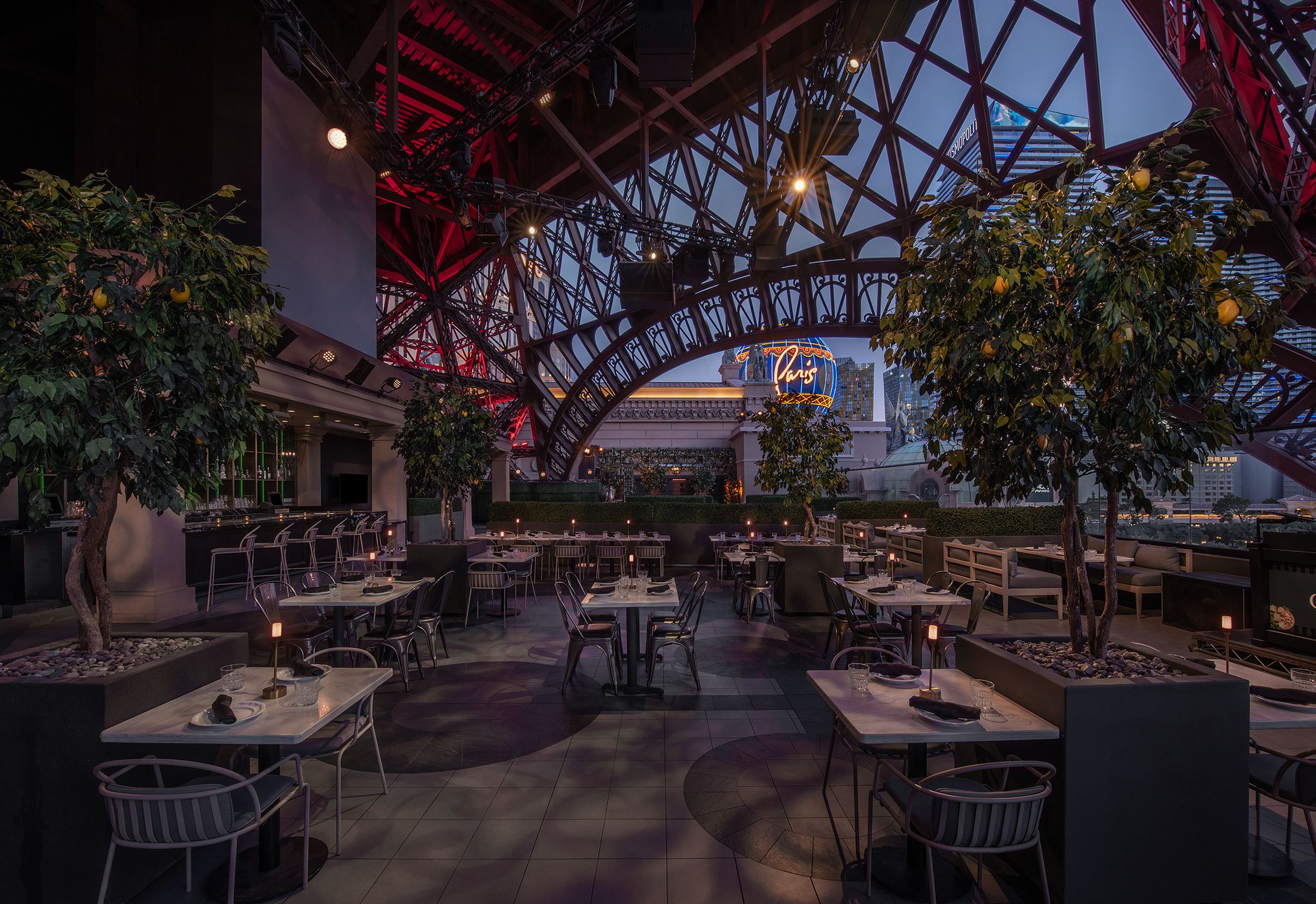 Cheri Rooftop dining area with tables, chairs, potted plants, and a view of Las Vegas at the base of the Eiffel Tower at Paris Las Vegas, illuminated by indoor lighting and sunset sky.