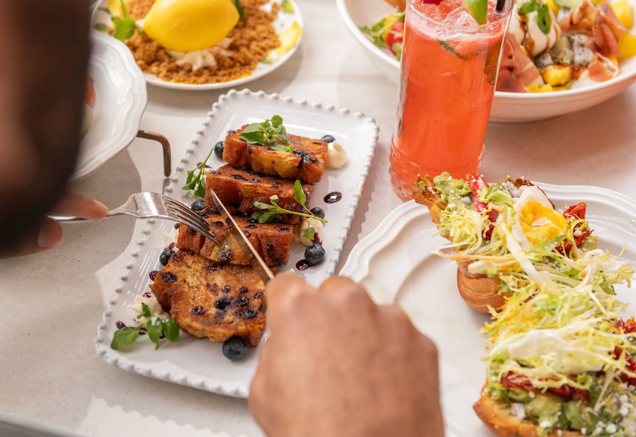 A person is cutting slices of blueberry bread with fresh blueberries and green herbs on top, surrounded by plated salads and a glass of pink beverage with ice and lime slices on a white table at Cheri Rooftop during brunch.