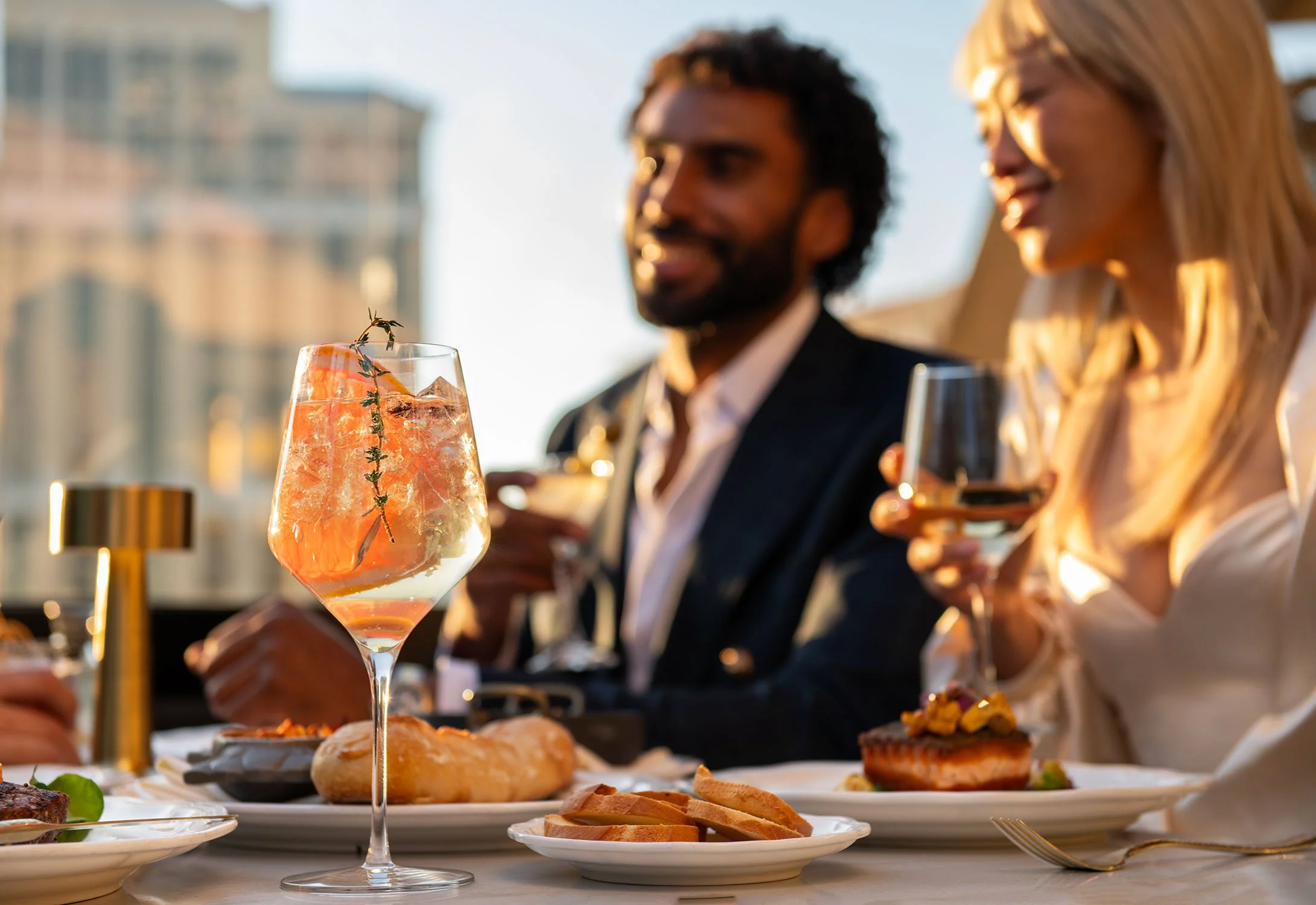 Two people enjoying dinner at Cheri Rooftop, with a focus on a glass of orange-colored cocktail garnished with a sprig of herbs.