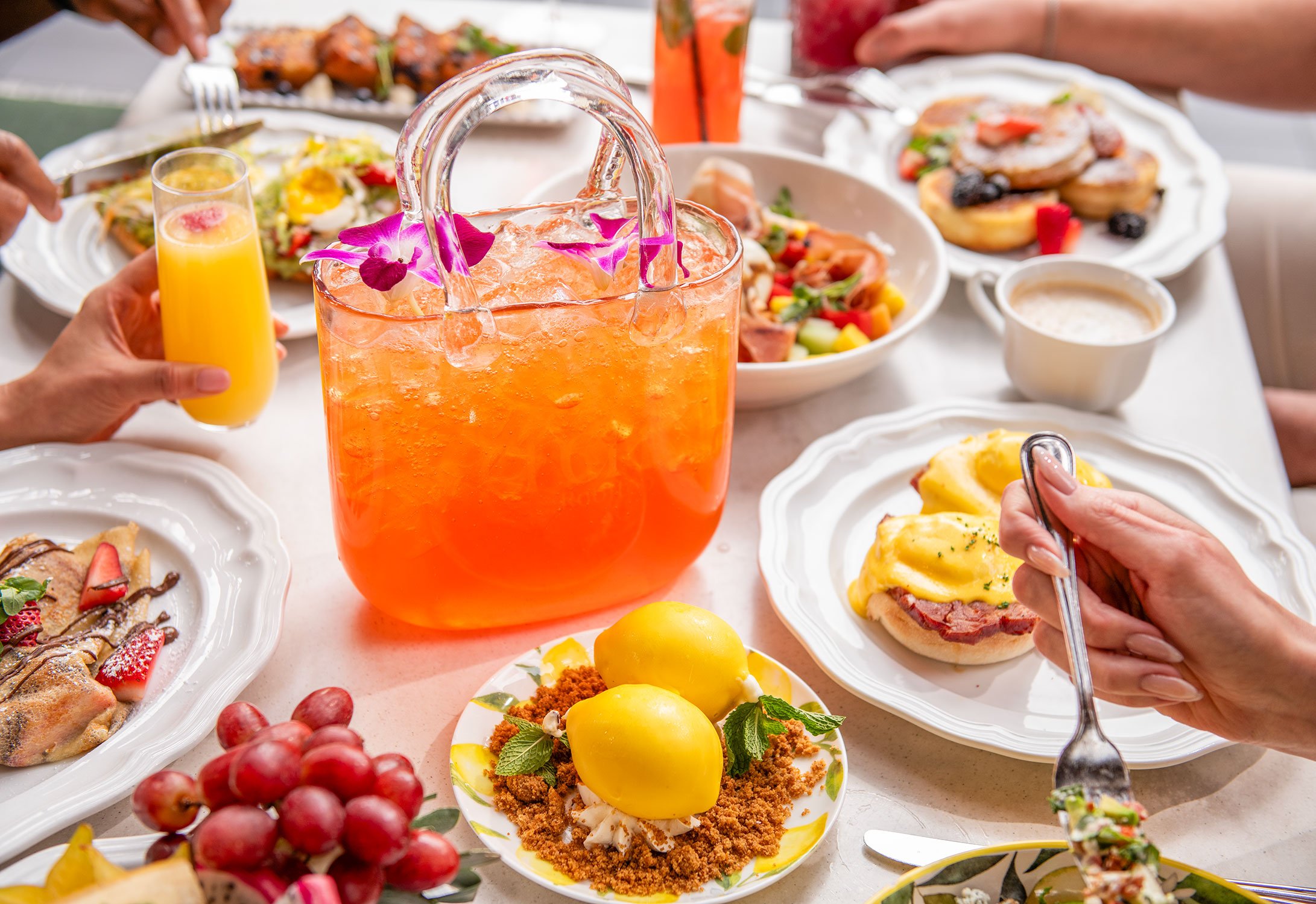 A table filled with various colorful brunch dishes including Cheri Rooftop's signature It Bag cocktail, fruit, salads, and desserts with people enjoying a meal.
