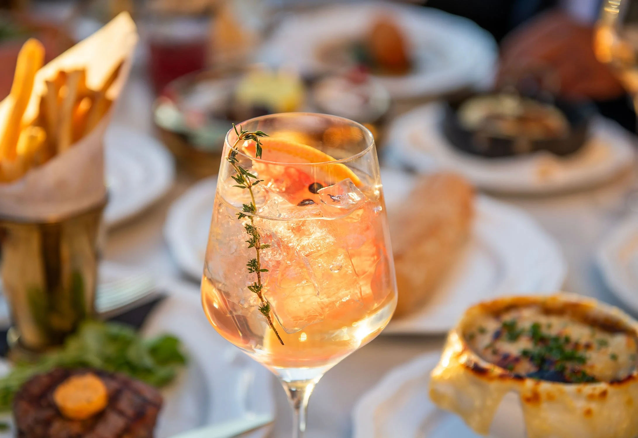A glass of pink cocktail garnished with a slice of orange and a sprig of thyme, with a blurred background of various appetizers and dishes on a table at Cheri Rooftop.
