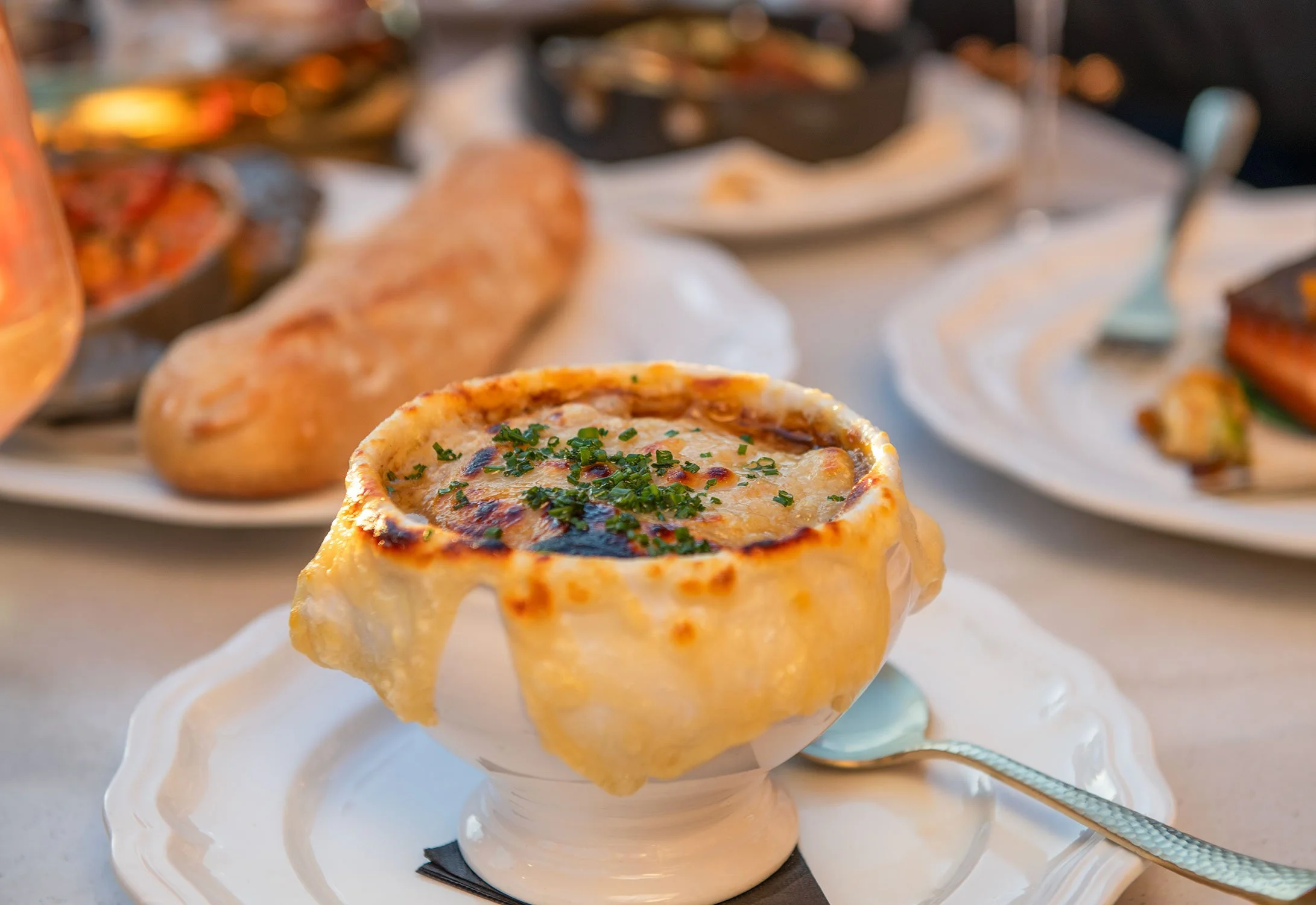 French Onion Soup in small round casserole topped with chopped herbs, with a creamy texture and sauce, placed on a white plate with a decorative edge in a restaurant setting with more dishes in the background  at Cheri Rooftop.