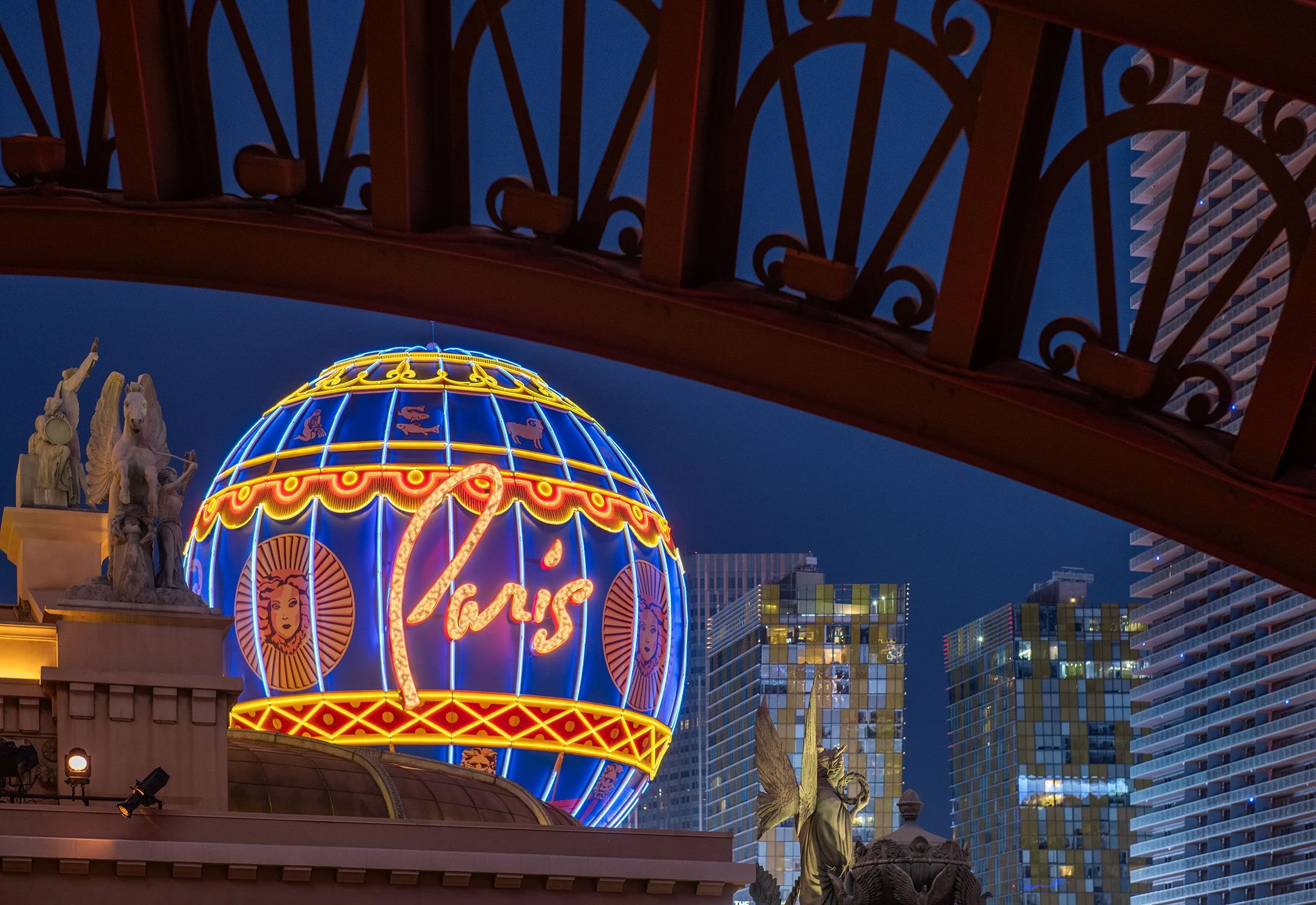 Night view of Las Vegas with the signature Paris ballon neon lit sign and decorative statues in the background, seen from Cheri Rooftop at the base of the Eiffel Tower at Paris Las Vegas.