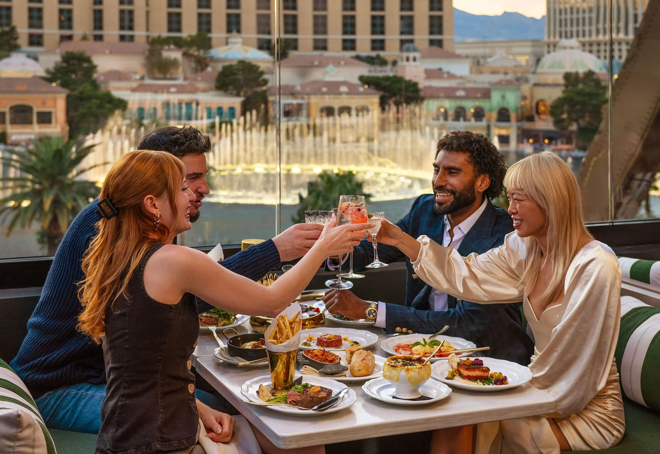 Group of four friends sit at a table outdoors, enjoying drinks and food at Cheri Rooftop with a Las Vegas Strip background including the Bellagio fountains.