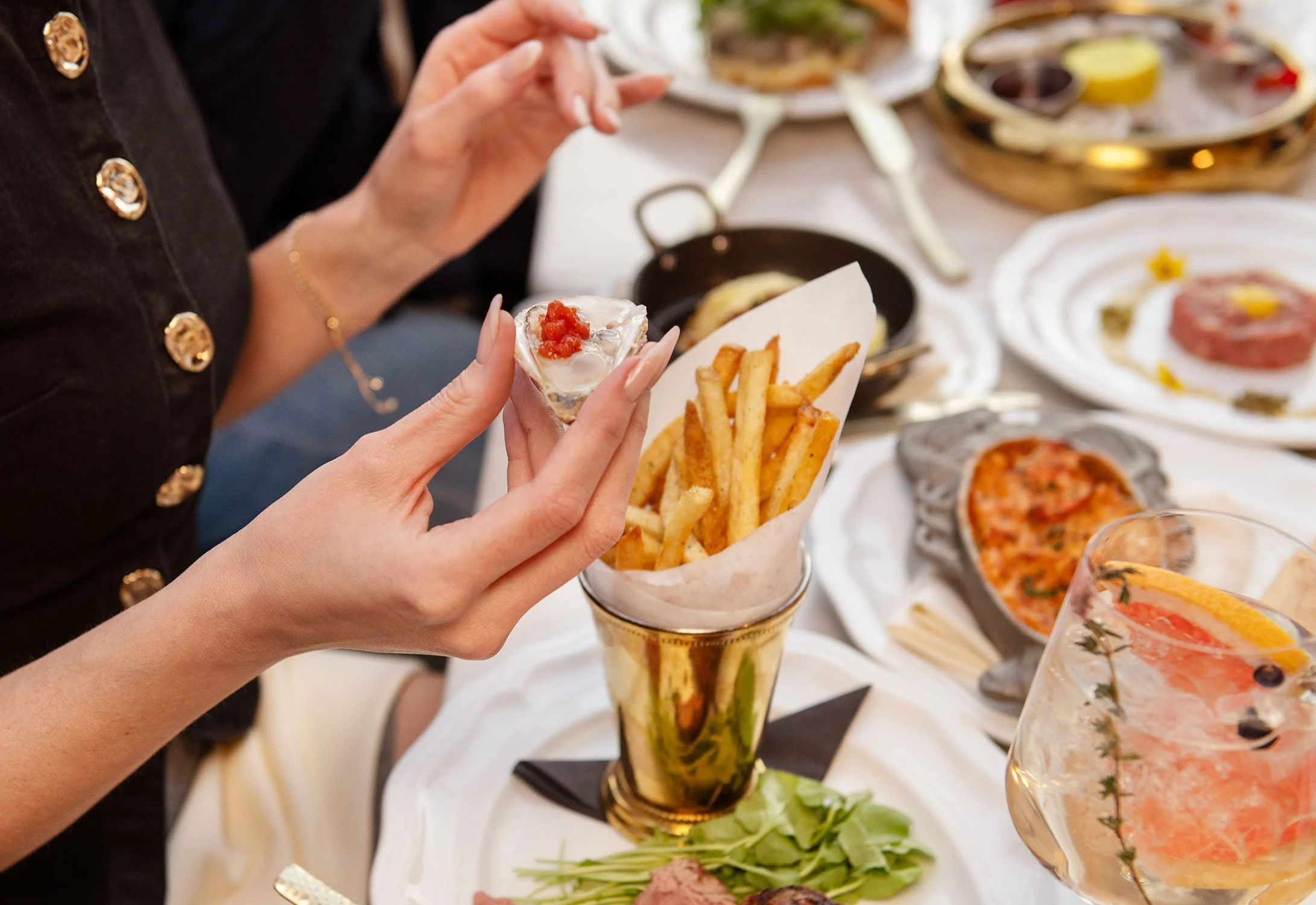 A person holding a small bowl of oyster topped with red sauce and a cone filled with French fries, with a table of food in the background at Cheri Rooftop.