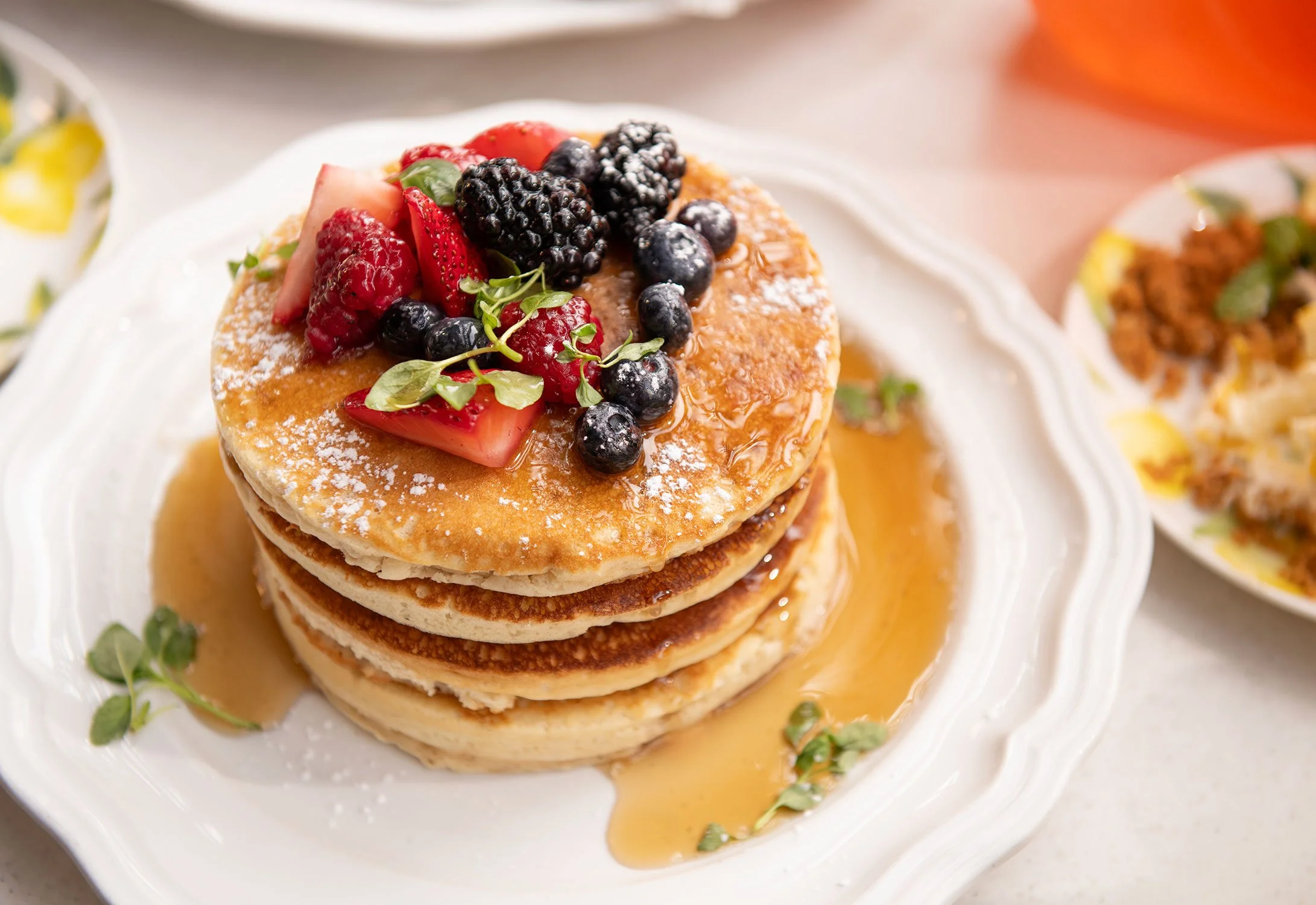 Stack of three golden brown pancakes topped with mixed berries and garnished with microgreens, drizzled with syrup on a white plate at Cheri Rooftop during brunch..