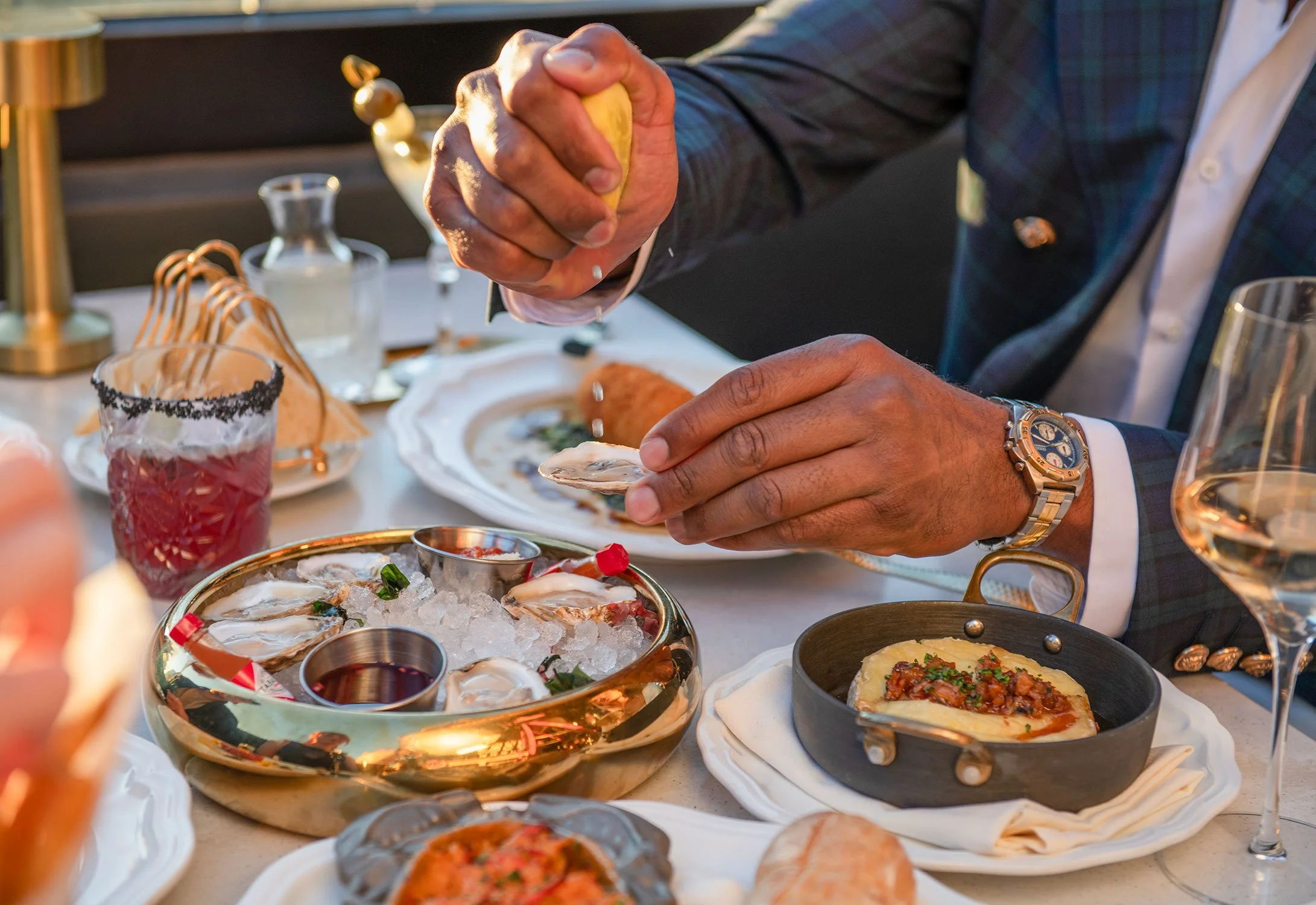 A person in a suit is enjoying various food items at Cheri Rooftop. The table has various dishes including oysters on ice, small bowls of condiments, and a skillet with a baked dish. There is a glass of red drink and a wine glass on the table.