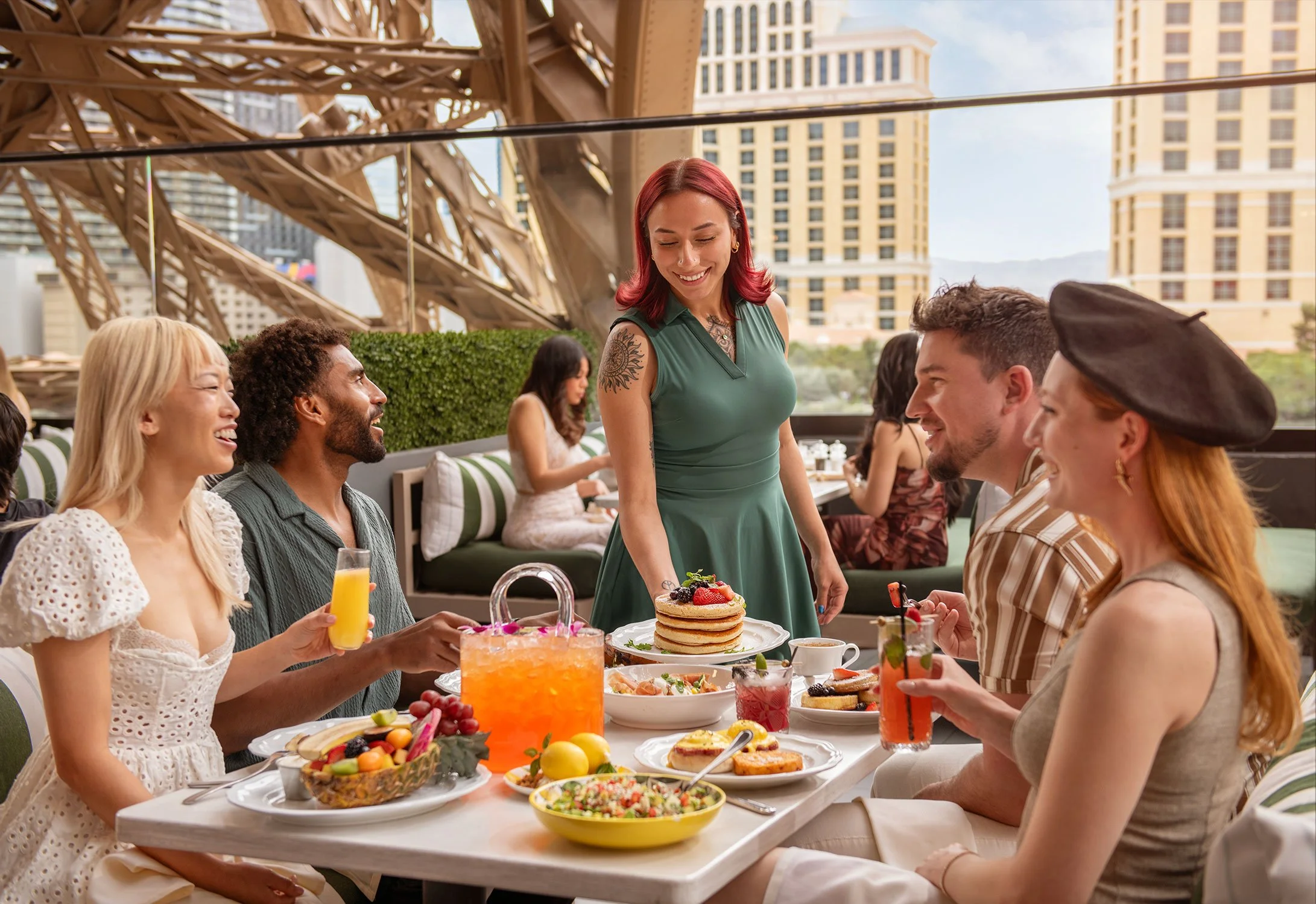 A group of friends enjoying a brunch at Cheri Rooftop with the base of Paris Las Vegas' Eiffel Tower and Bellagio Resort in the background. A woman is serving pancakes, while others are holding drinks and conversing.