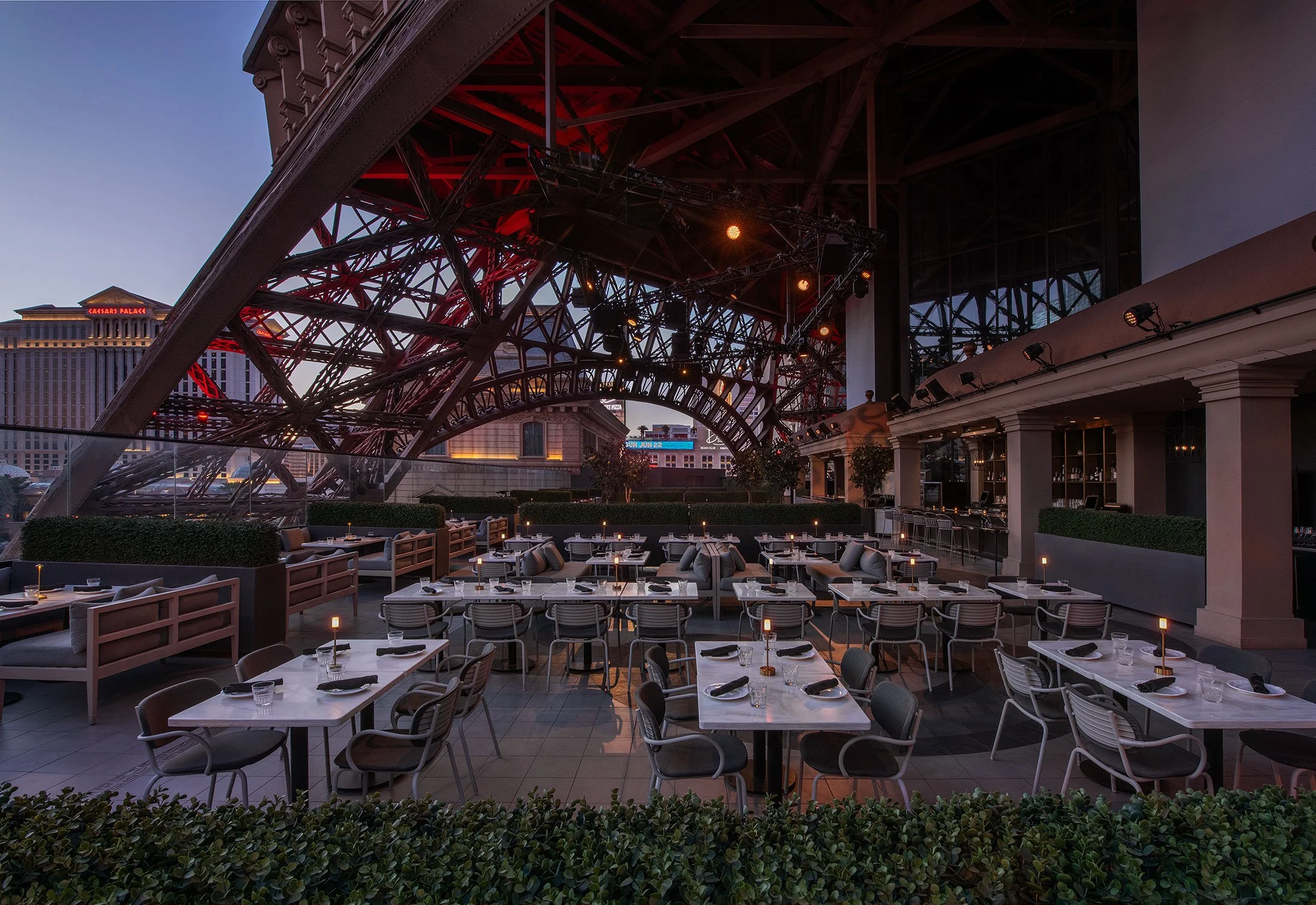 Cheri Rooftop's dining area with tables, chairs, and candles under the base of the Eiffel Tower at Paris Las Vegas  during golden hour, with the Las Vegas Strip in the background.