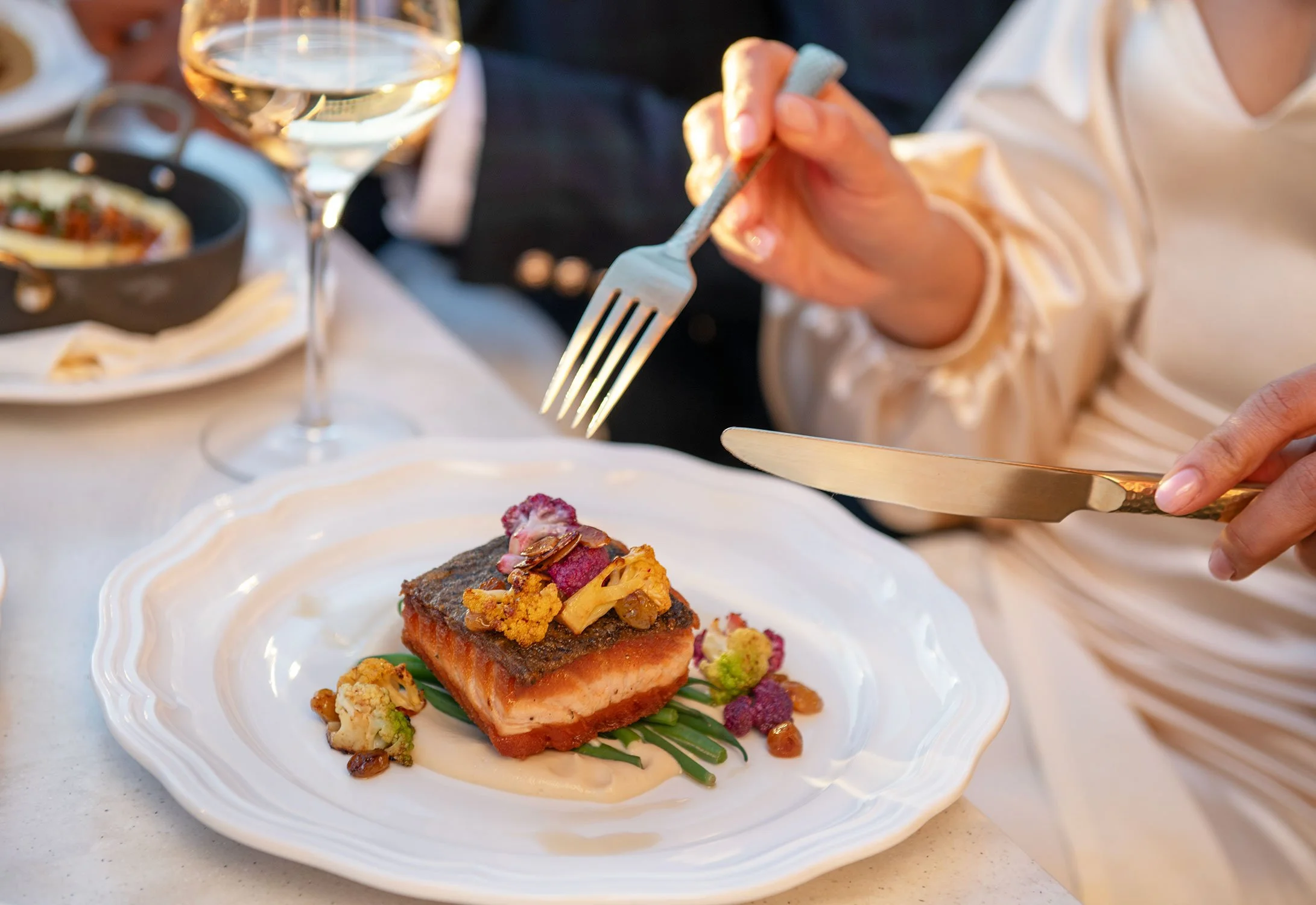 Close-up of a plated gourmet dish with a piece of seared salmon topped with garnish, served with a cream sauce and vegetables, on a white ornate plate. A person in a cream-colored shirt uses a fork and knife to cut the dish, with a glass of white win