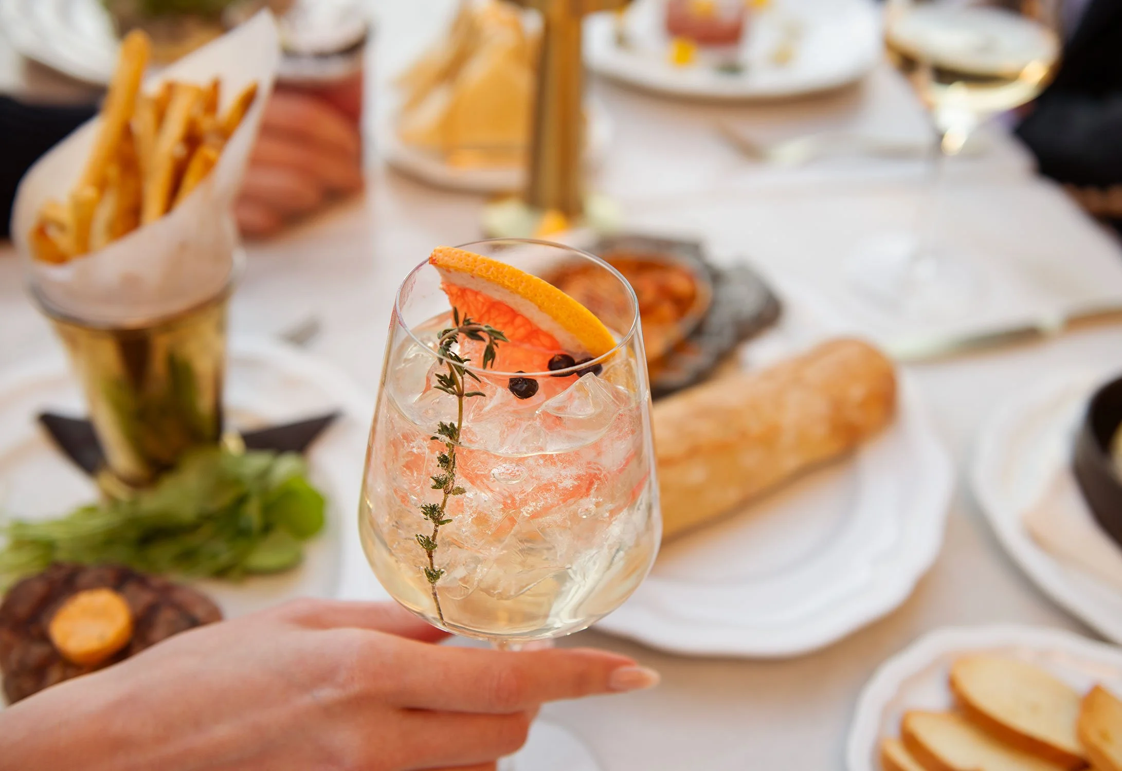 A hand holding a glass with a cocktail garnished with an orange slice, a sprig of thyme, and small dark berries, with a table of food and drinks in the background at Cheri Rooftop.