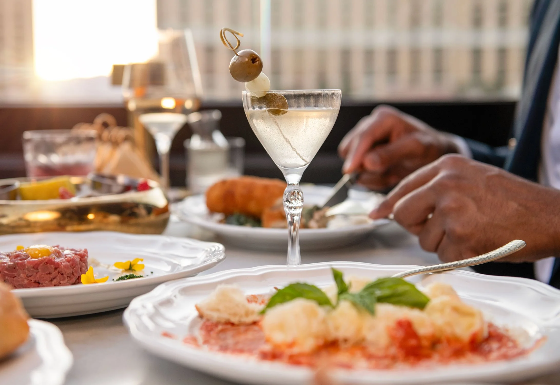 A martini glass with a garnish of olives and an onion, set on a table with plates of food in the background at a dining setting  at Cheri Rooftop.