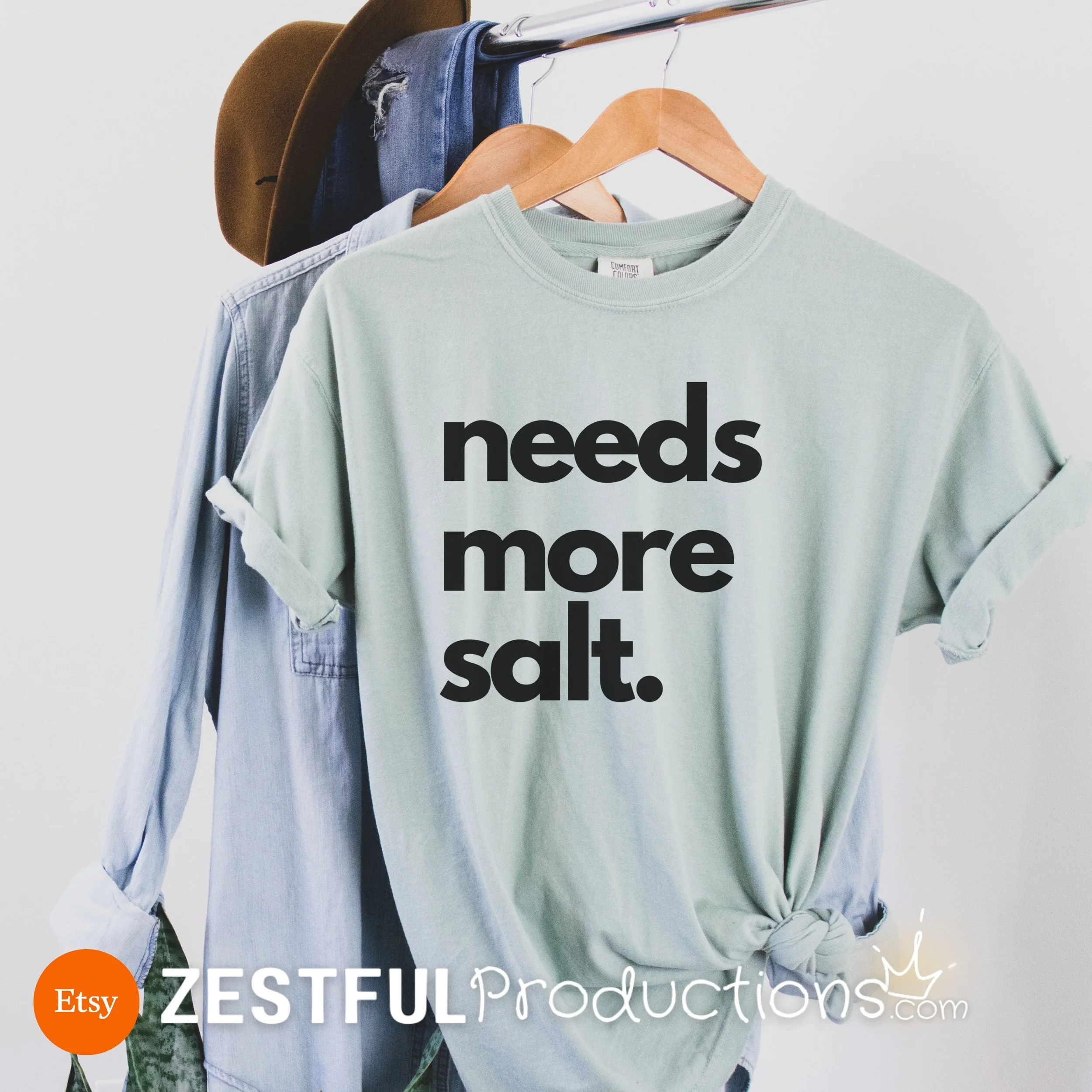 Bay Green Comfort Colors shirt with bold black 'Needs More Salt' text, hanging on a rack – sarcastic foodie gift tee.