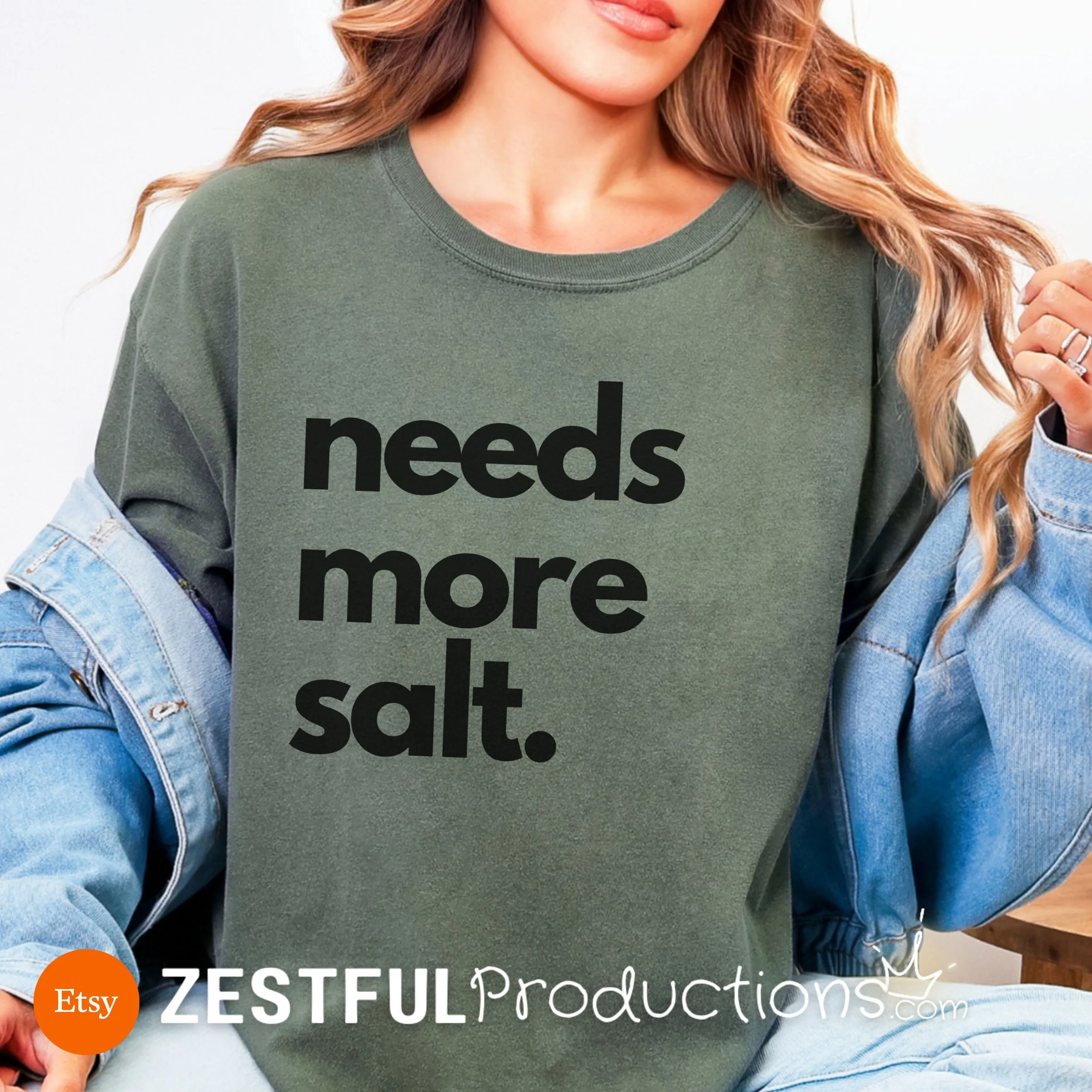 Woman in cozy moss green Comfort Colors shirt with black ‘Needs More Salt’ quote – bold foodie kitchen humor tee.