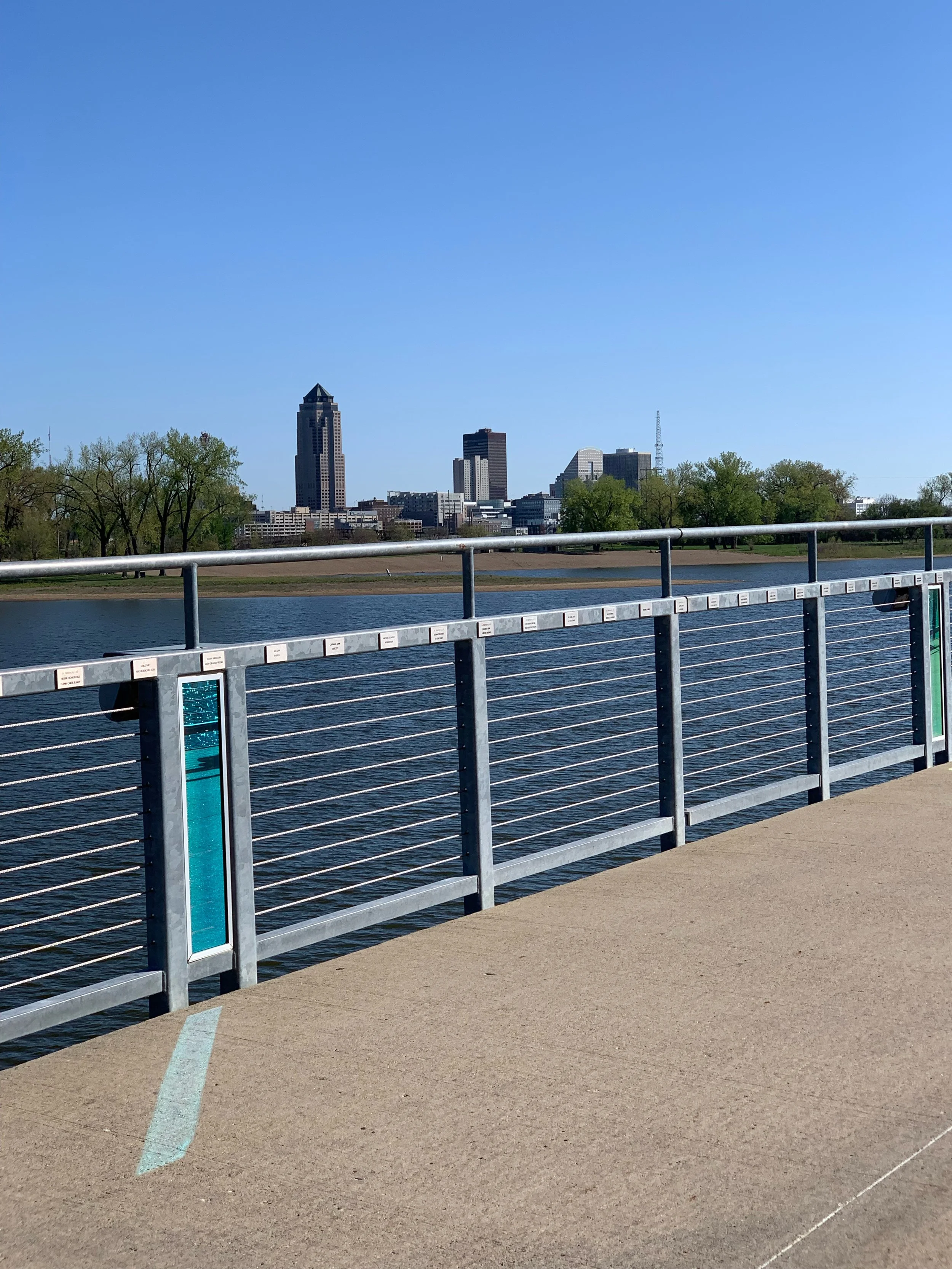 City skyline with tall buildings across a river, viewed from a paved walkway with a metal railing.