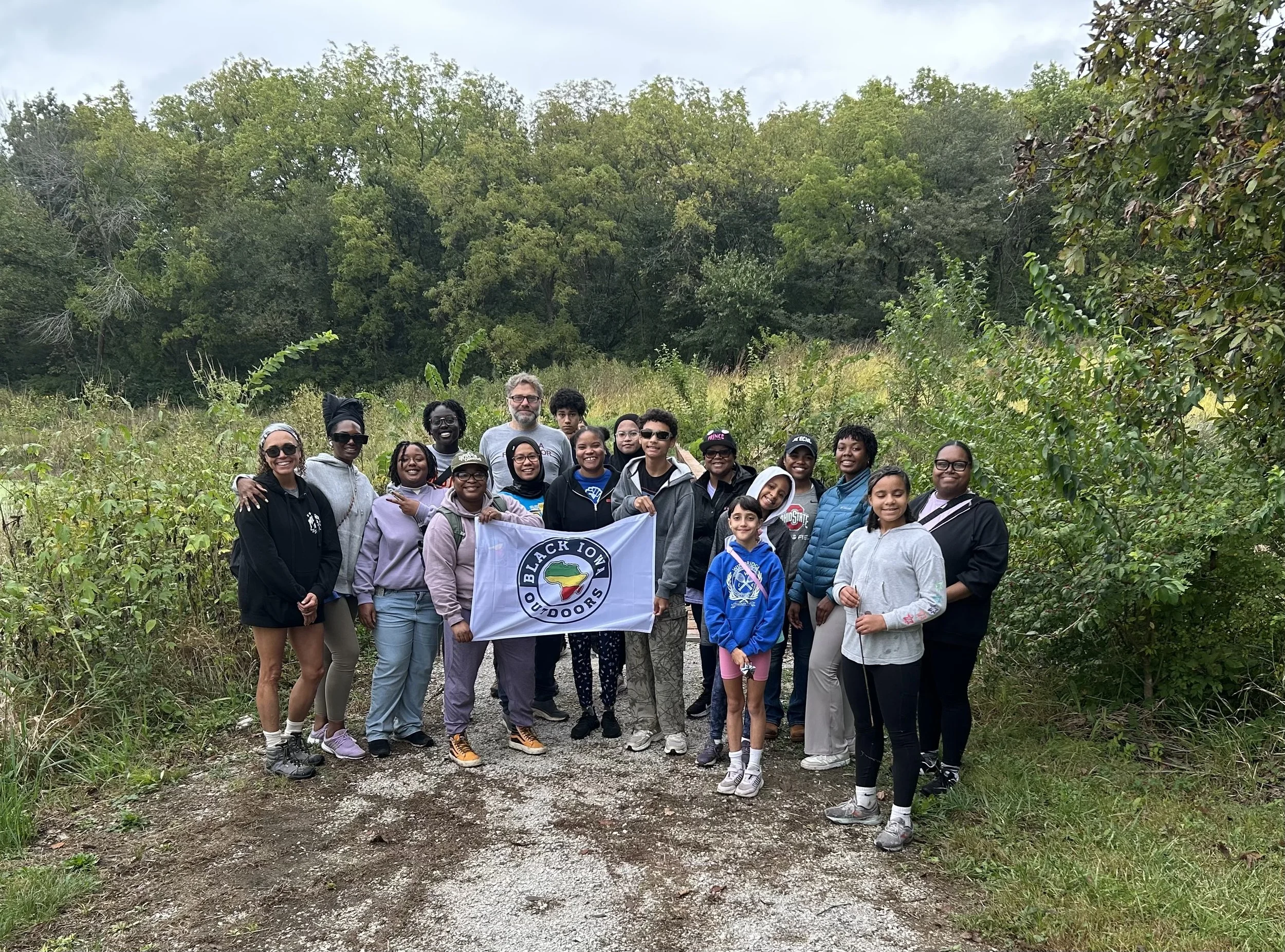 Group of diverse people outdoors holding a 'Black Iowa Outdoors' flag, standing on a dirt trail surrounded by green trees and bushes