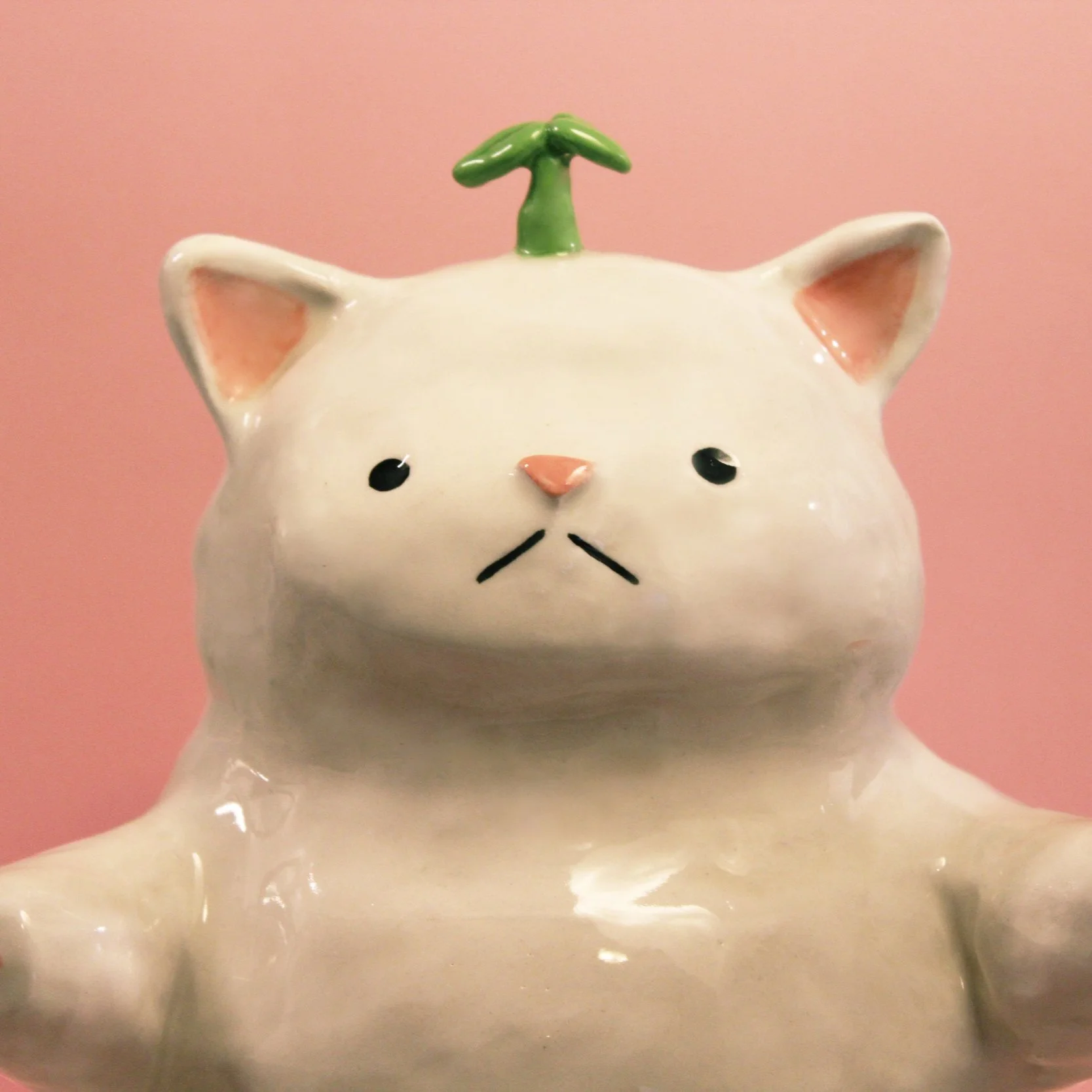 Ceramic cat figurine with a pink background, a green sprout on its head, and a simple, cute face.