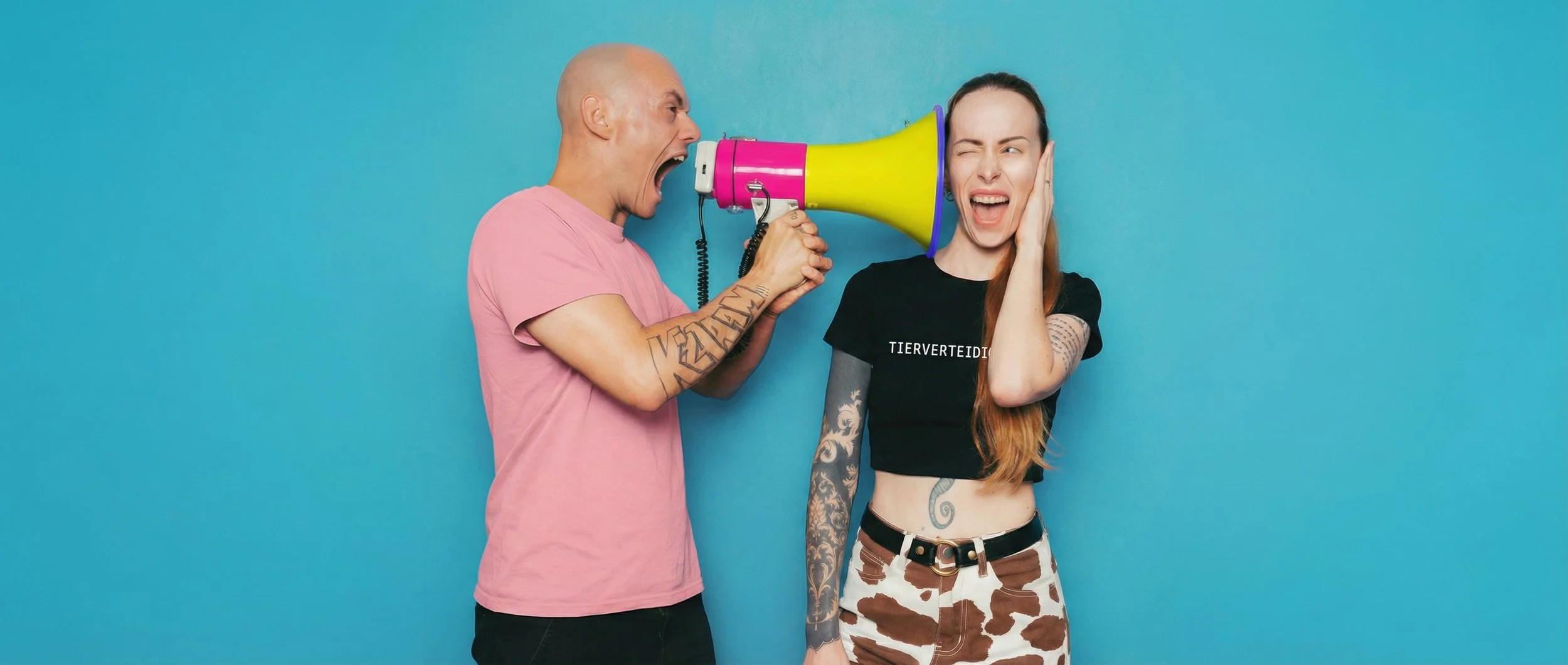 Ein Mann schreit durch eine bunte Megaphon, während eine Frau am Ohr hält und lachend reagiert, vor einem blauen Hintergrund.