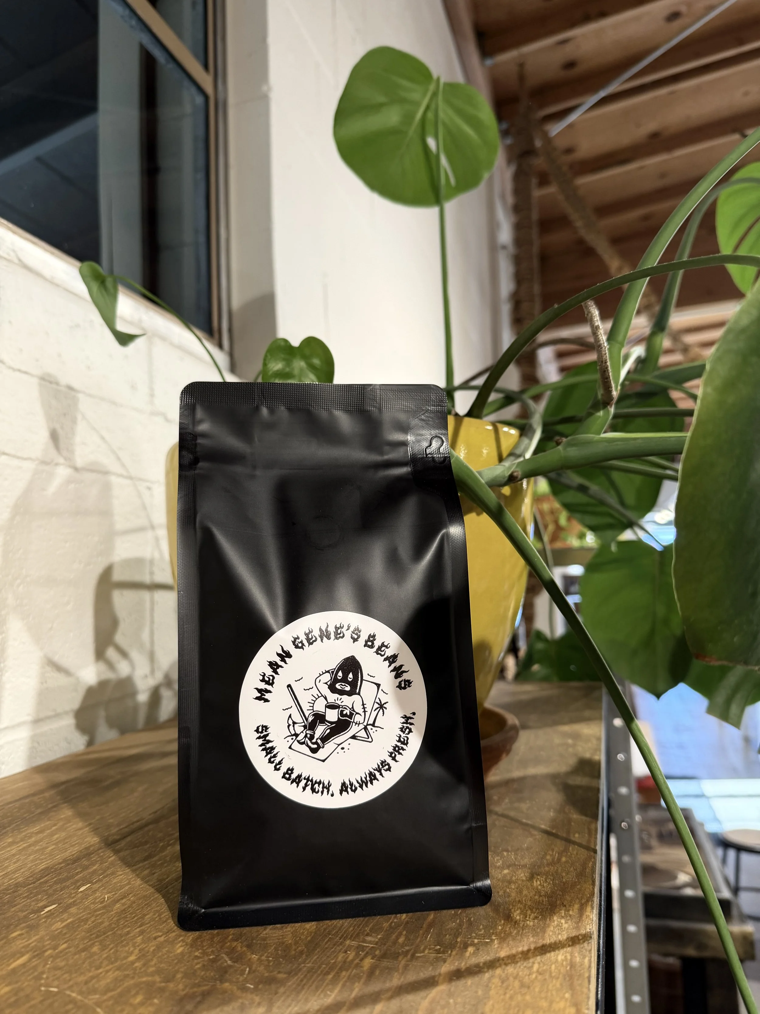 A black bag with a white circular sticker featuring a cartoon woman holding a coffee cup, surrounded by text, placed on a wooden surface with green plants and a window in the background.