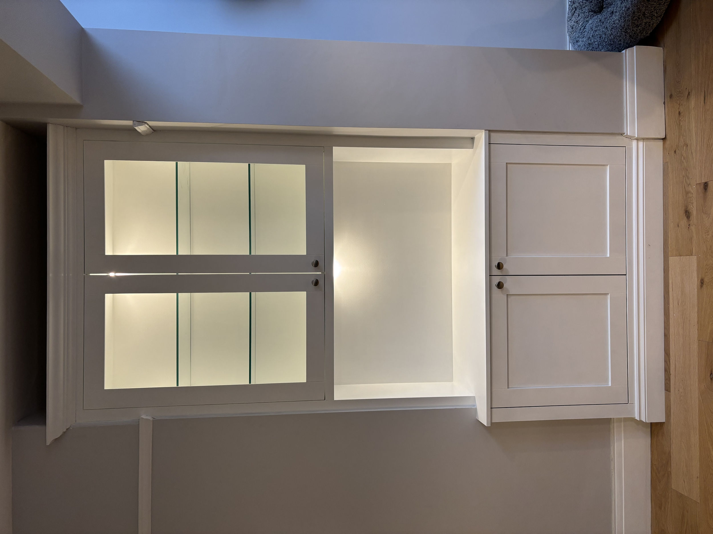 A white display cabinet with glass doors and drawers, located in a room with wooden flooring and a gray wall.