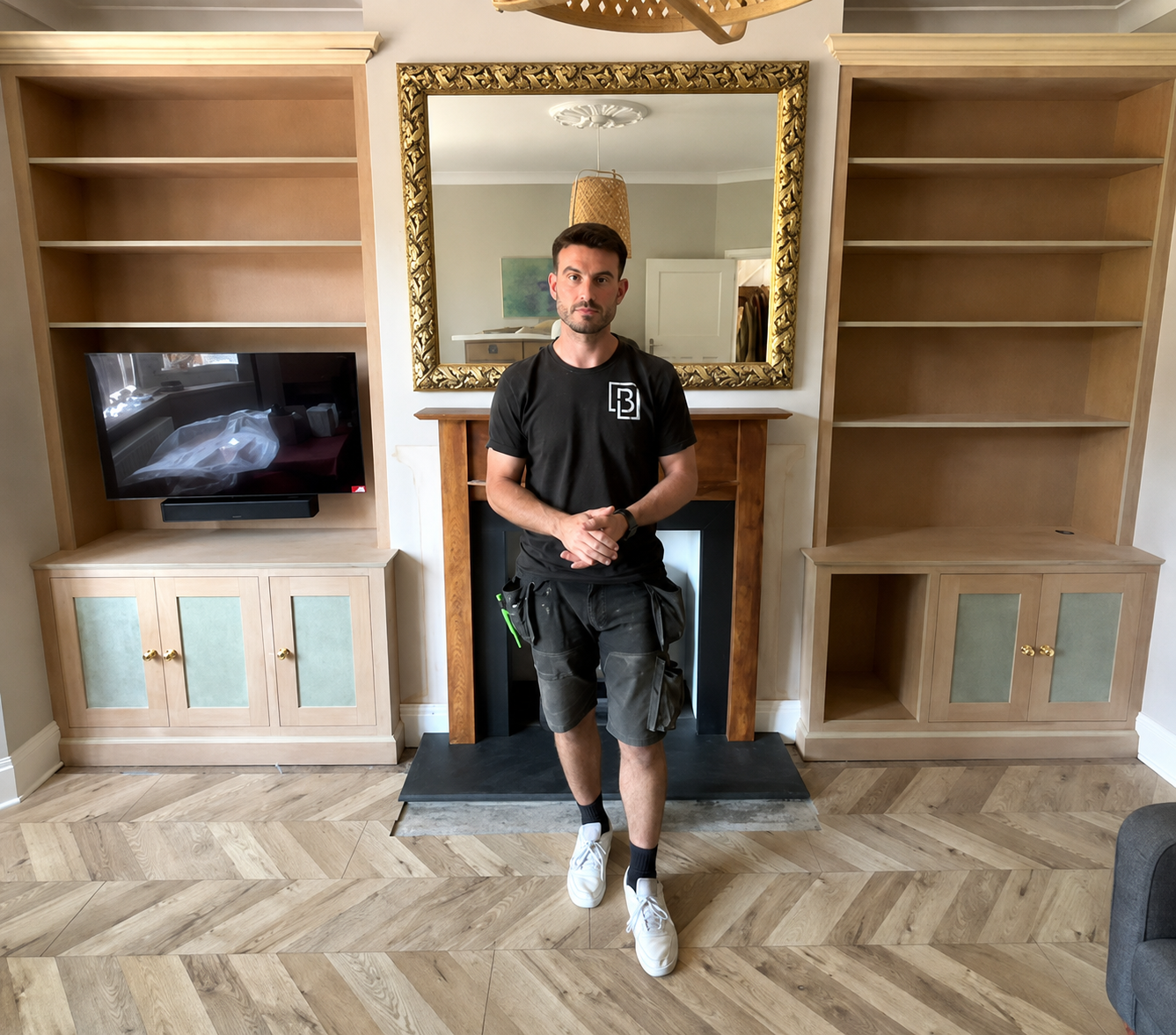 A young man stands indoors in front of a decorative mirror and fireplace, flanked by empty built-in shelves, wearing a black t-shirt, shorts, and white sneakers.