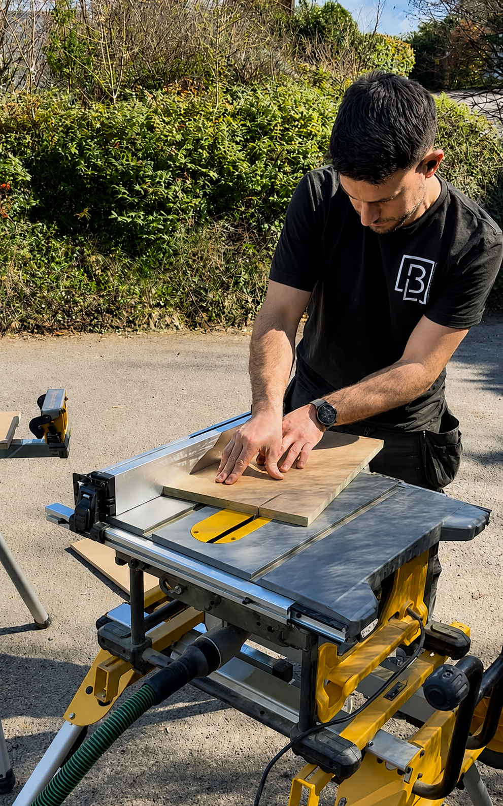 Man working on a woodworking project outdoors using a table saw to cut a wooden board. He is wearing a black shirt and a wristwatch, with green bushes and trees in the background.
