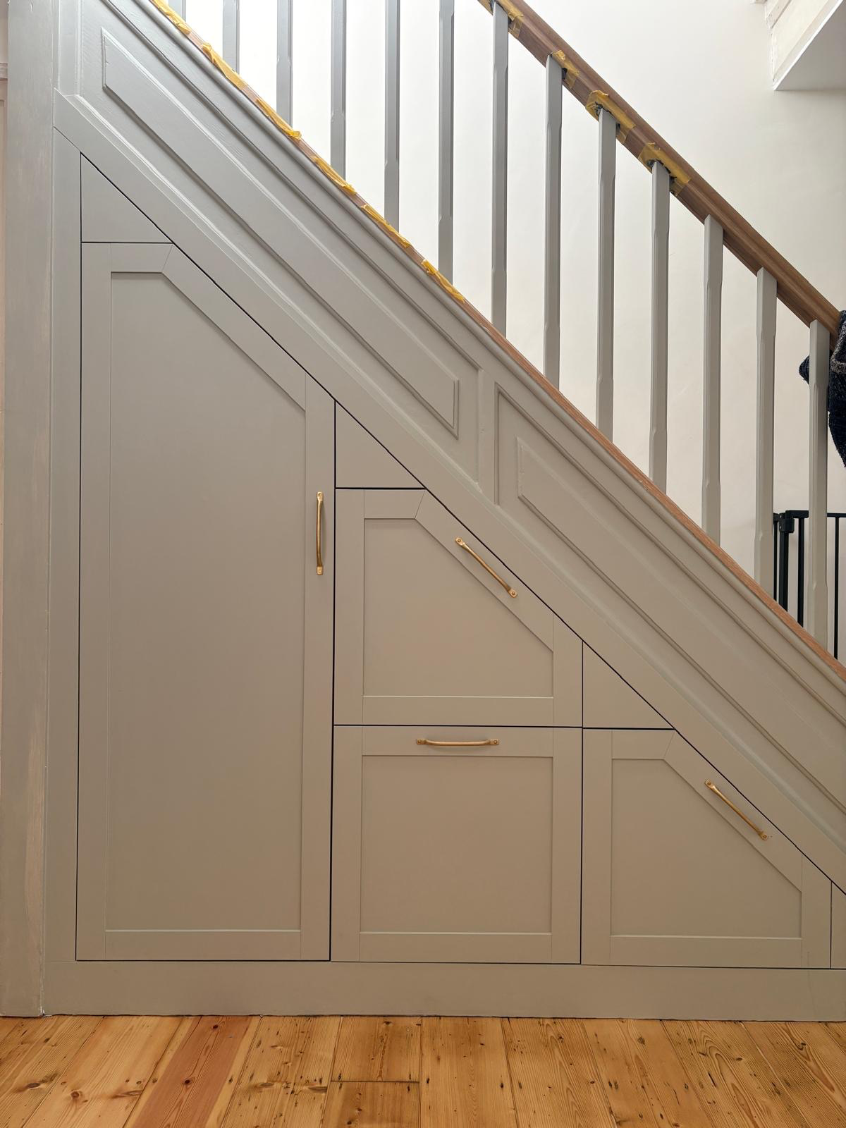 Under-stairs storage with cabinetry and gold handles in a home, with a staircase railing and balusters above and wooden flooring below.