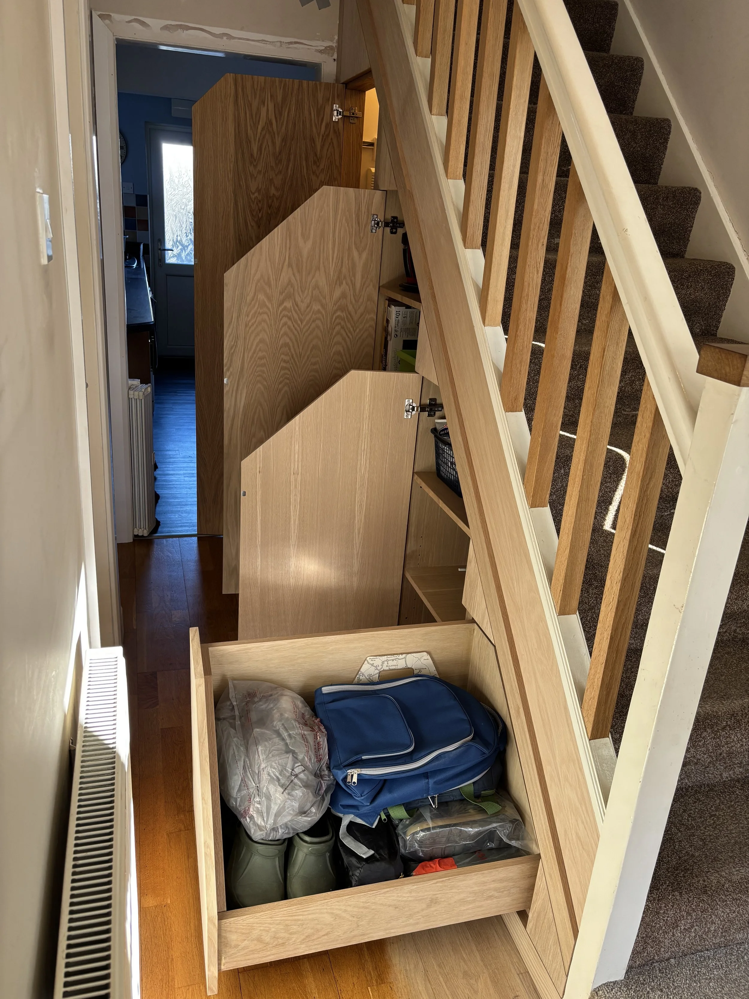 Empty wooden staircase storage space filled with various items including a blue duffel bag, a plastic bag, and green watering cans.