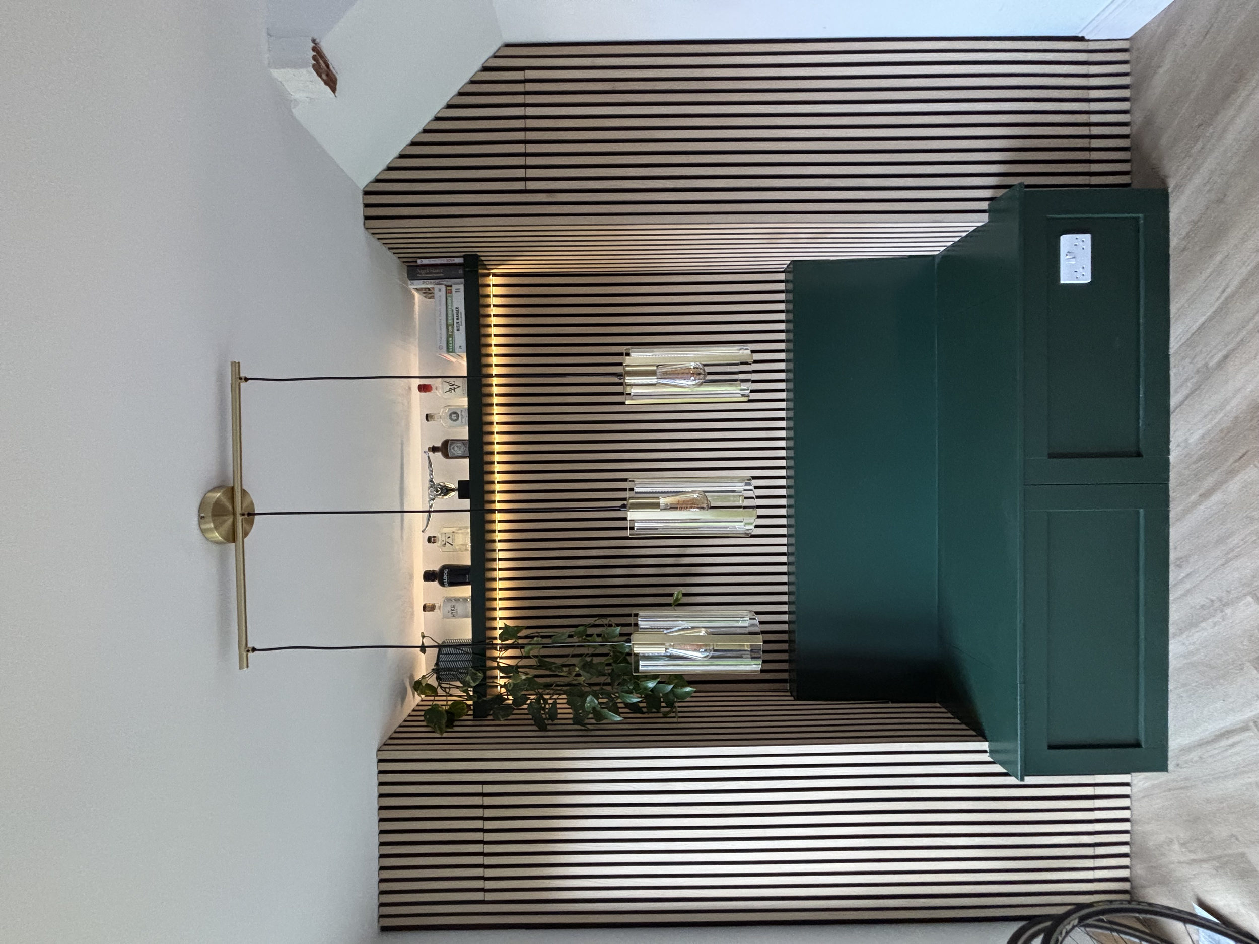 Modern interior with a black and white striped wood accent wall, a dark green console table with glass vases, a gold light fixture, potted plants, and a floating shelf with books.