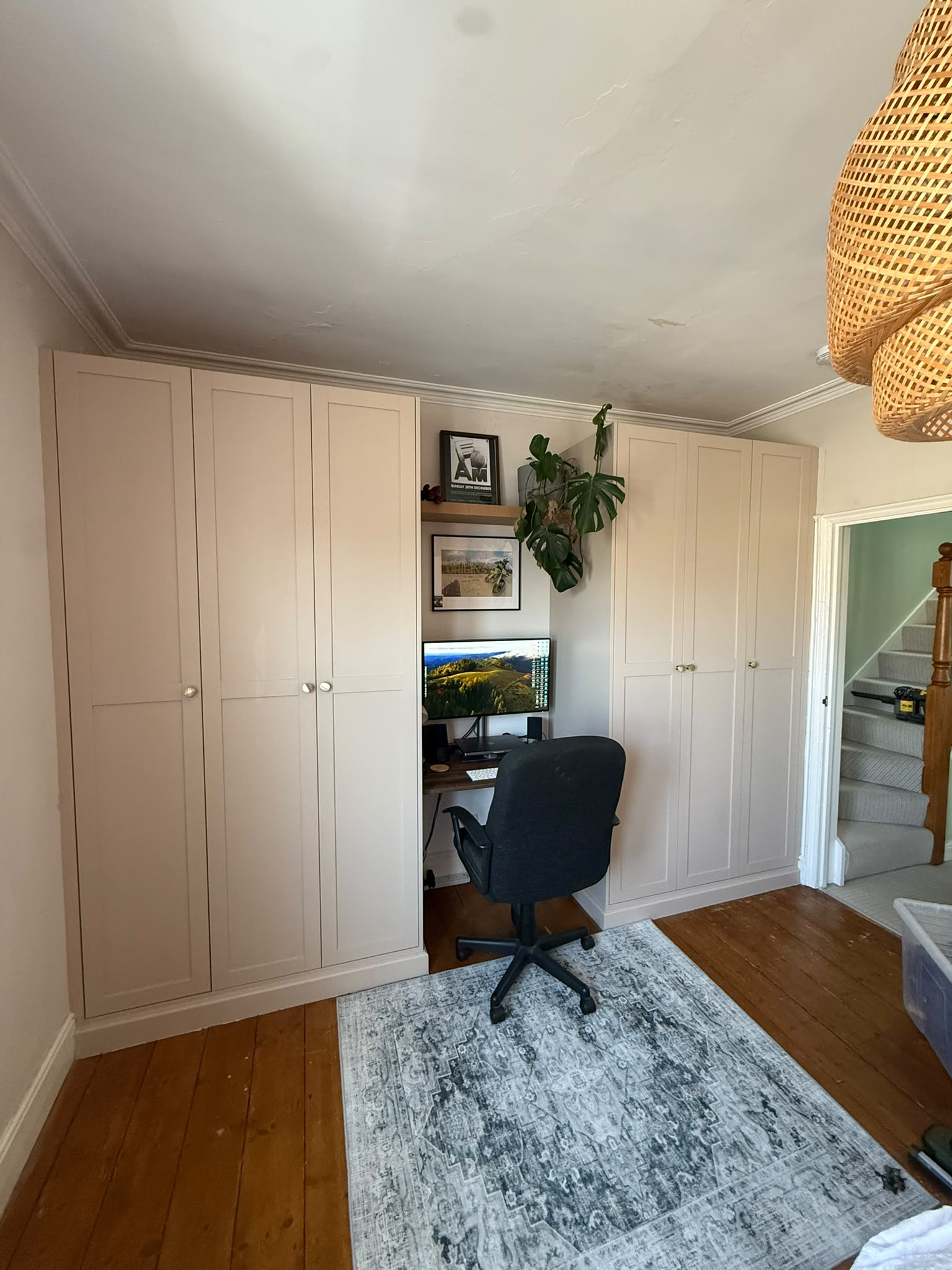 Home office corner with wardrobe, desk, black office chair, framed pictures, green plant, flatscreen monitor, and a staircase nearby.