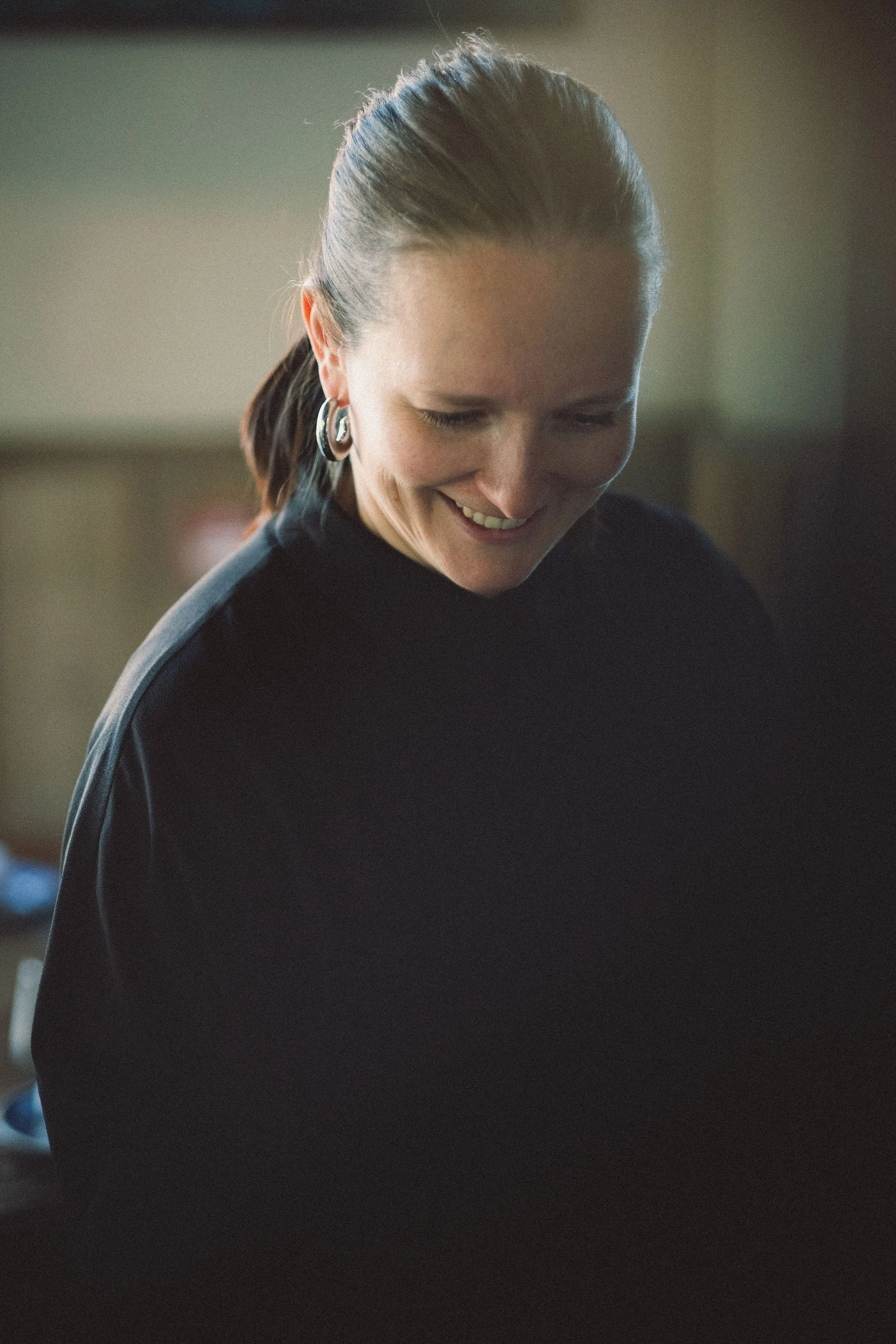 Close-up van lachende vrouw met bruin haar in een staart, draagt grote zilveren oorringen en een zwarte trui, binnen.