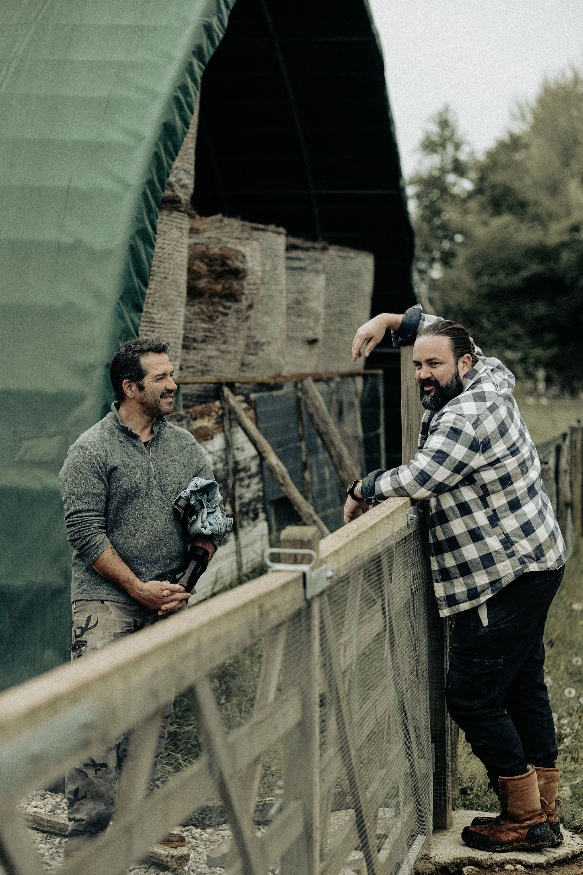 Twee mannen praten bij een hek op een boerderij, achtergrond met schuurruimtes en bomen.