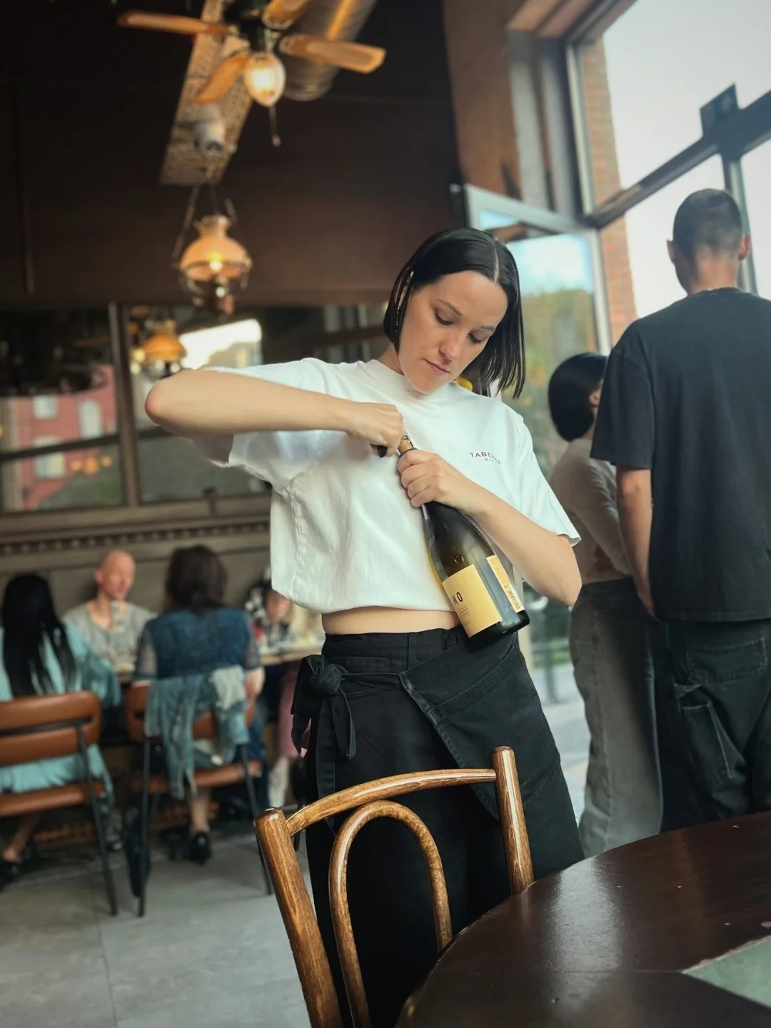 Vrouw opent een fles wijn in een restaurant met andere gasten op de achtergrond en grote ramen die natuurlijk licht binnenlaten.