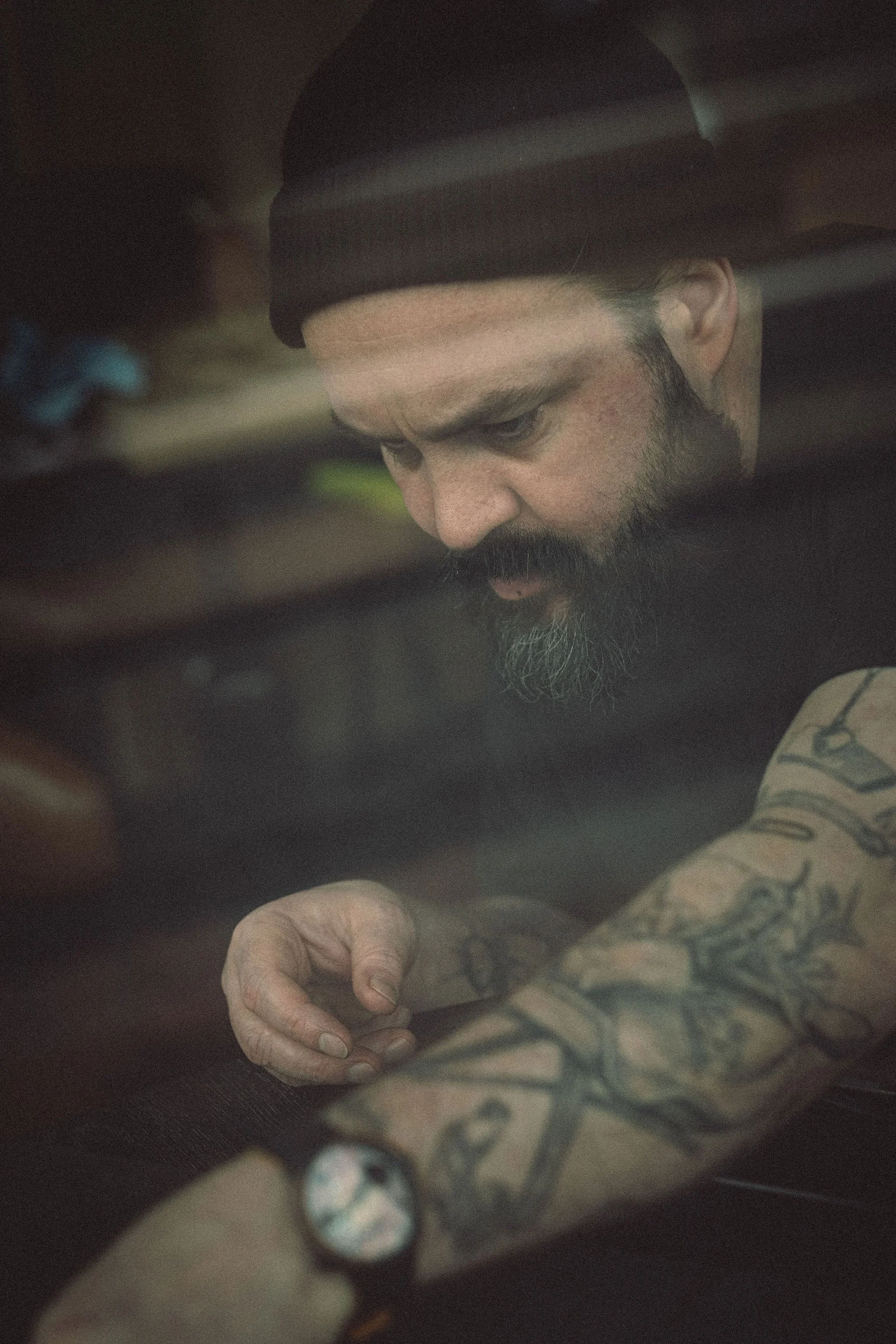 Een man met een baard, getatoeëerde arm en een zwart horloge, die zich door een raam of reflectie kijkt.