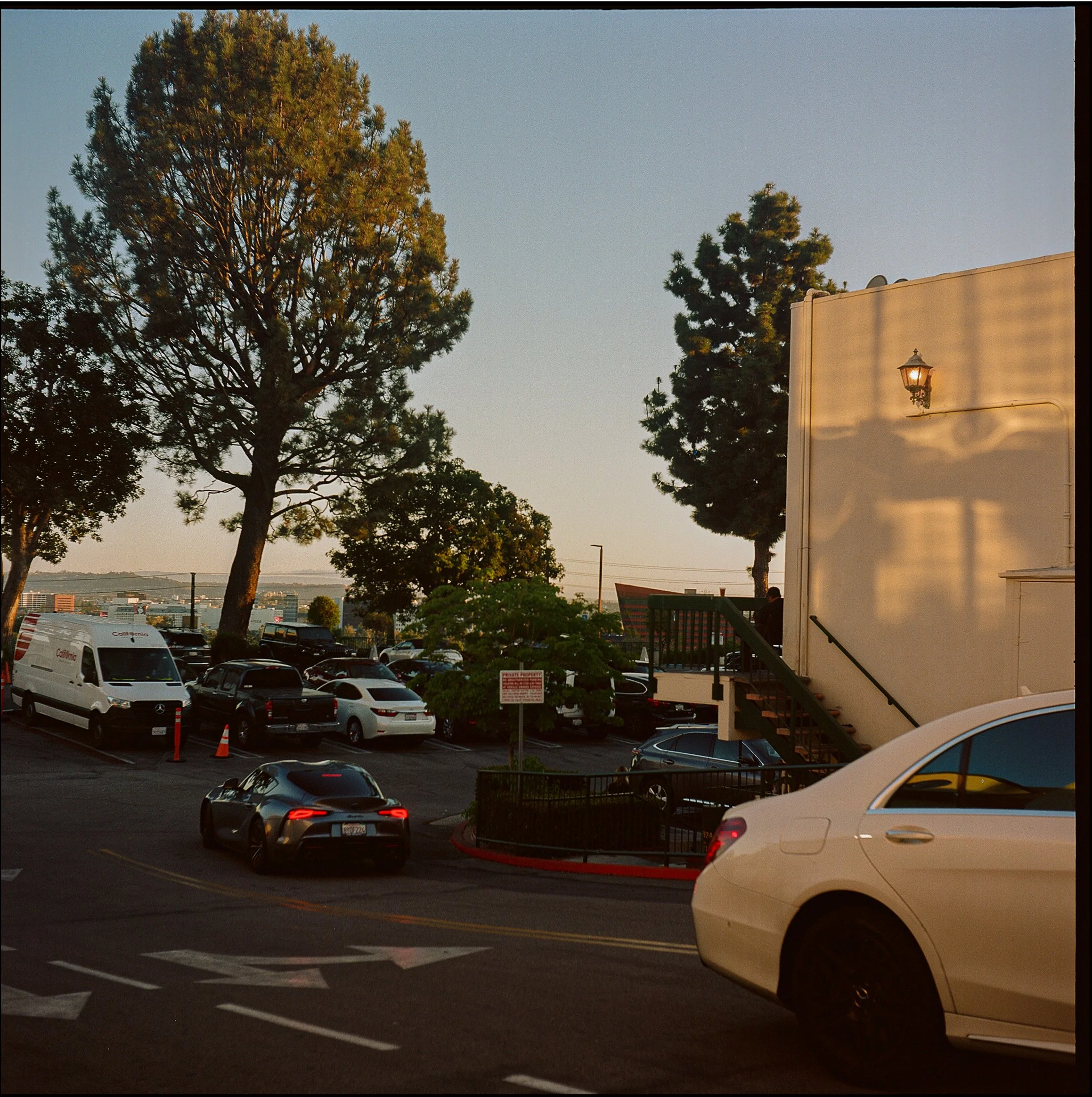 Parking lot with several cars, trees, a building with an outdoor light, and a view of a cityscape and hills in the distance during sunset.