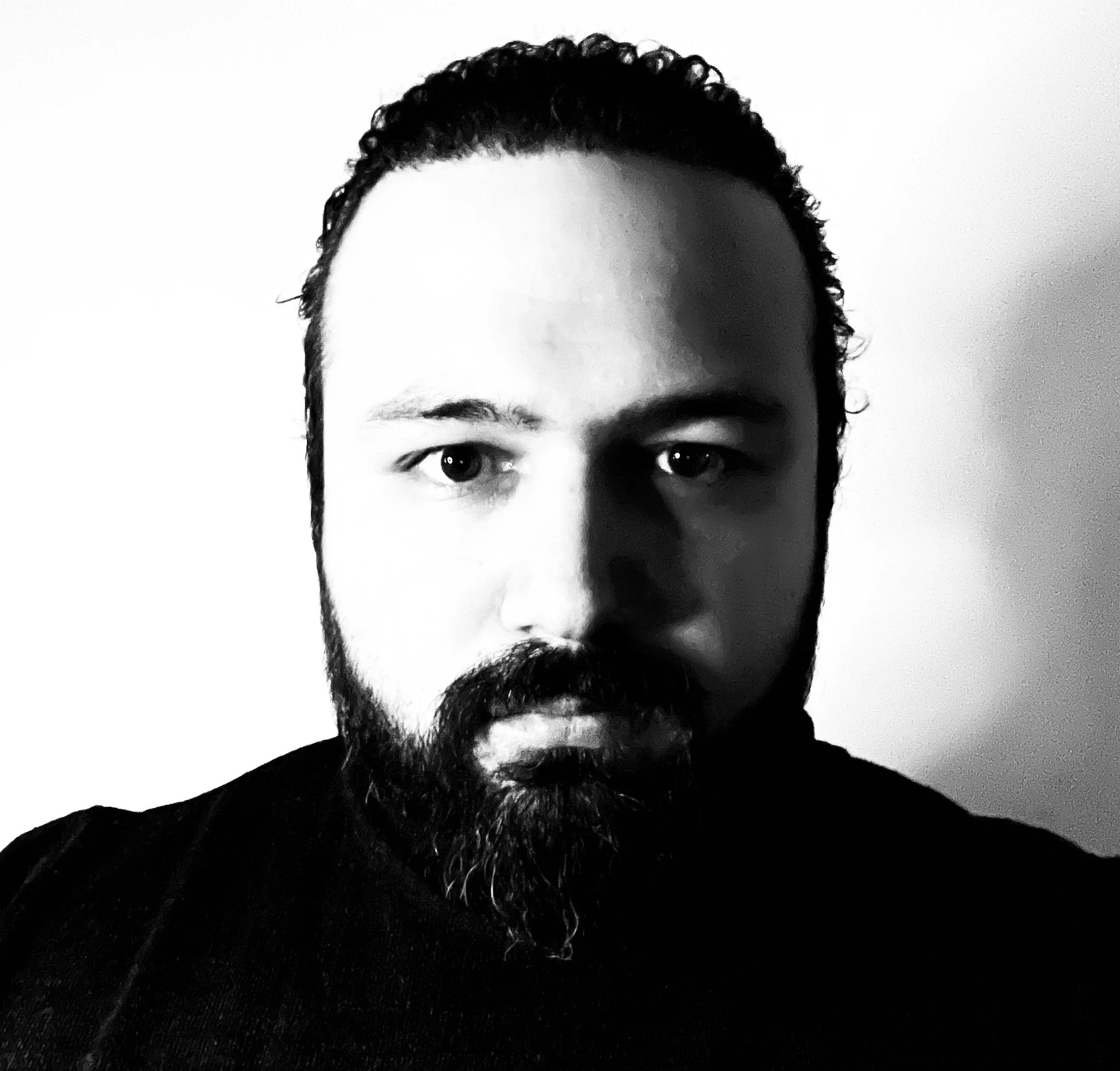 Black and white close-up photo of a man's face with a beard and short hair, looking directly at the camera.