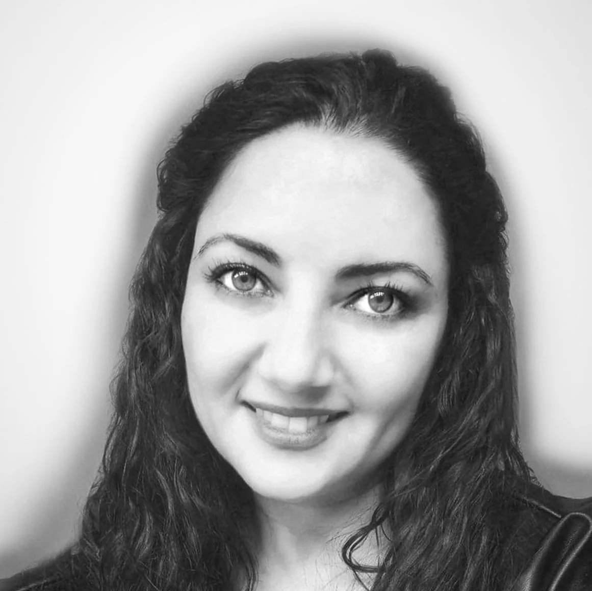 Black and white portrait of a woman with long, curly hair and light-colored eyes, smiling slightly, wearing a dark top.