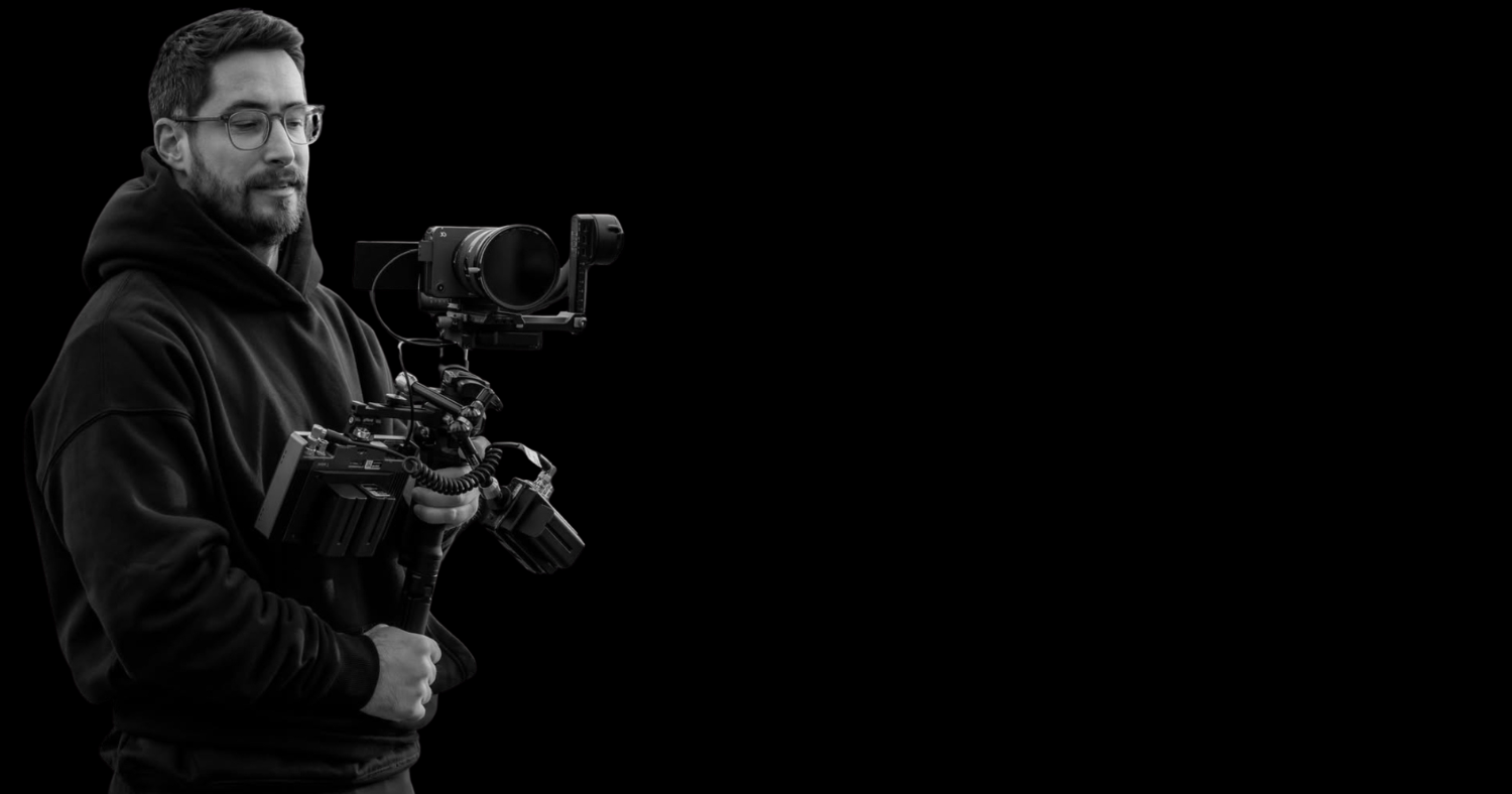 Black and white photo of a man holding a professional video camera on a gimbal, wearing glasses and a hoodie, against a plain black background.
