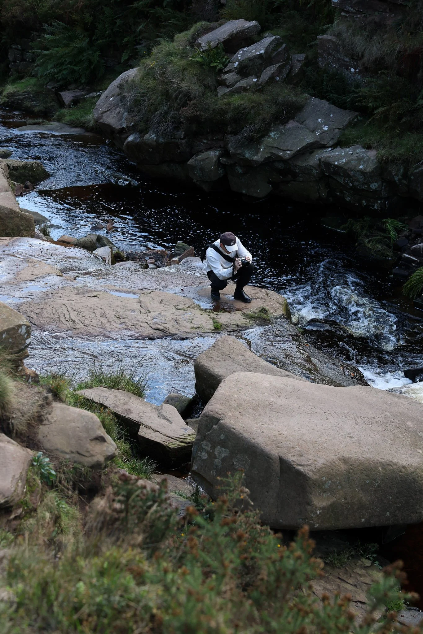 A person crouching on a large rock in a river, holding a camera, surrounded by large rocks and lush greenery.