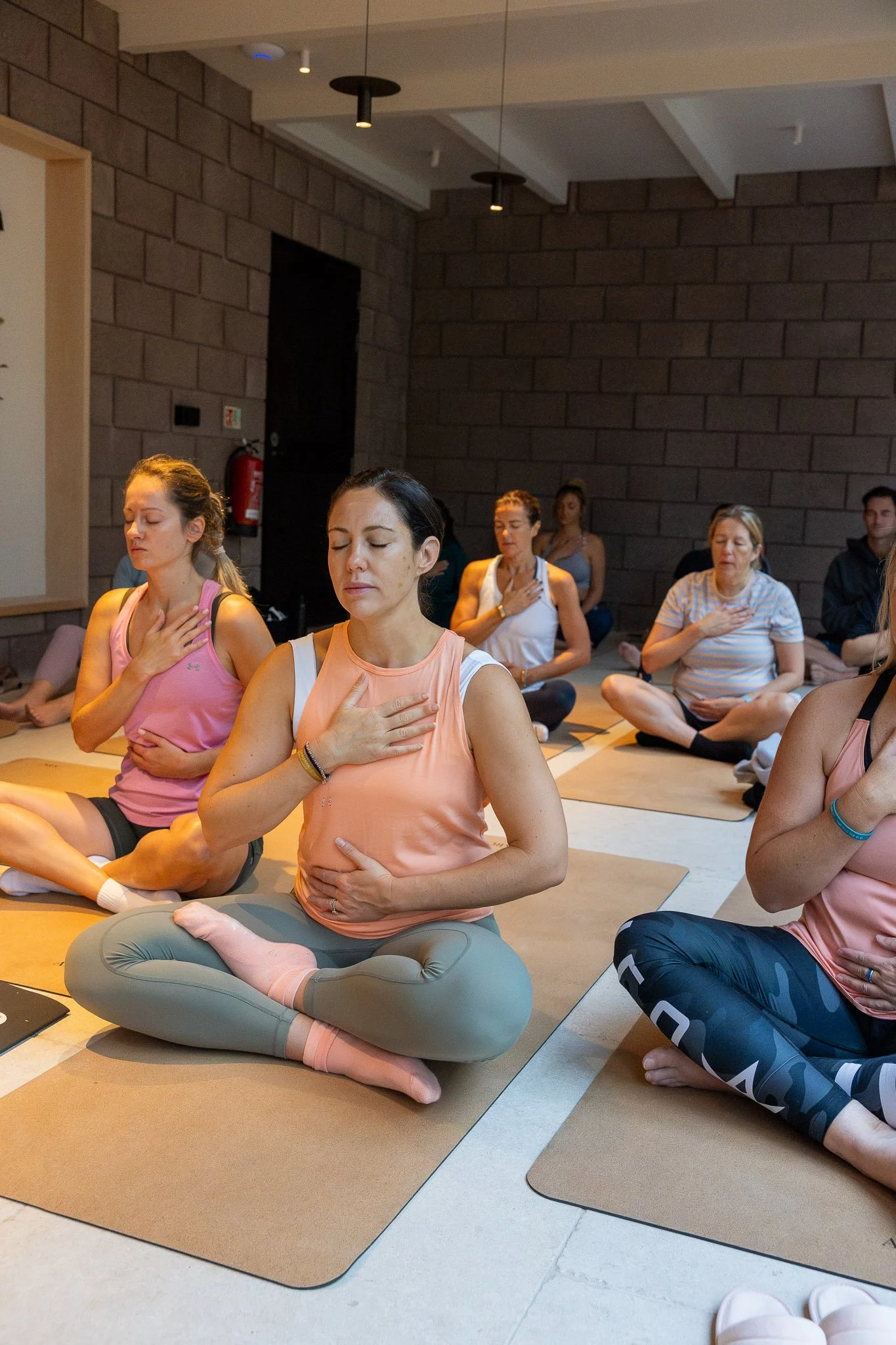 Group of women participating in a meditation or mindfulness class, sitting cross-legged on mats with hands over their hearts and eyes closed, in a room with dark brick walls.