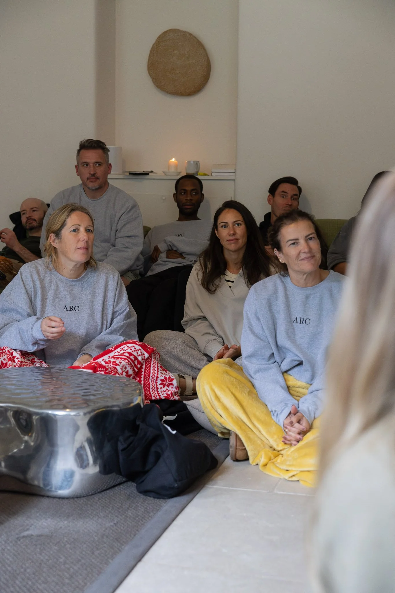 Group of people sitting on the floor and sofa, watching something, with some wearing gray sweatshirts with 'ARC' printed on them, casual clothing, and blankets around.