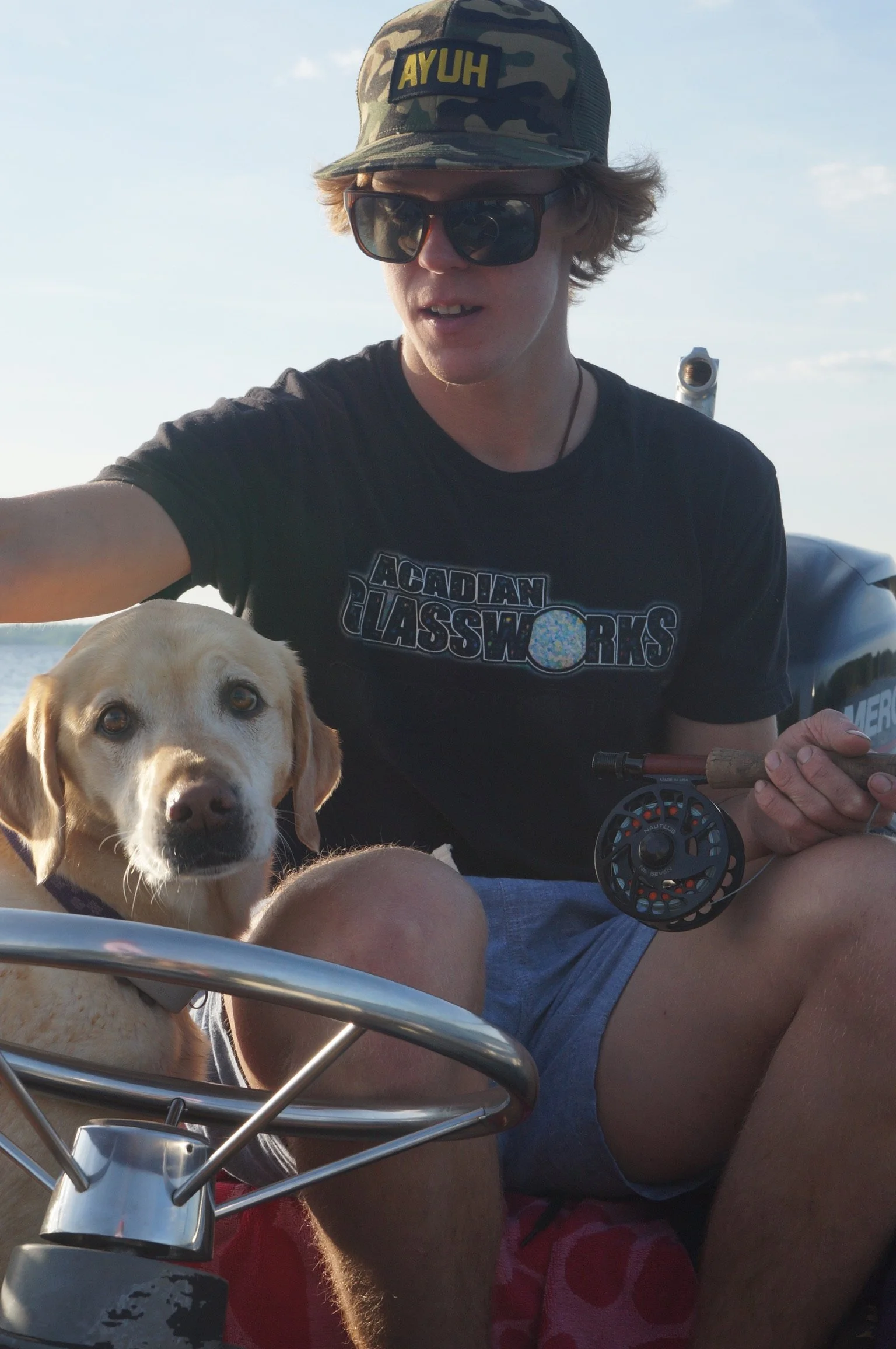 A young man wearing sunglasses, a camouflage cap with the word 'AYUH' on it, and a black t-shirt with 'ACADIAN CLASSWORKS' printed on it, sitting outdoors on a boat with a dog, holding a fishing rod.
