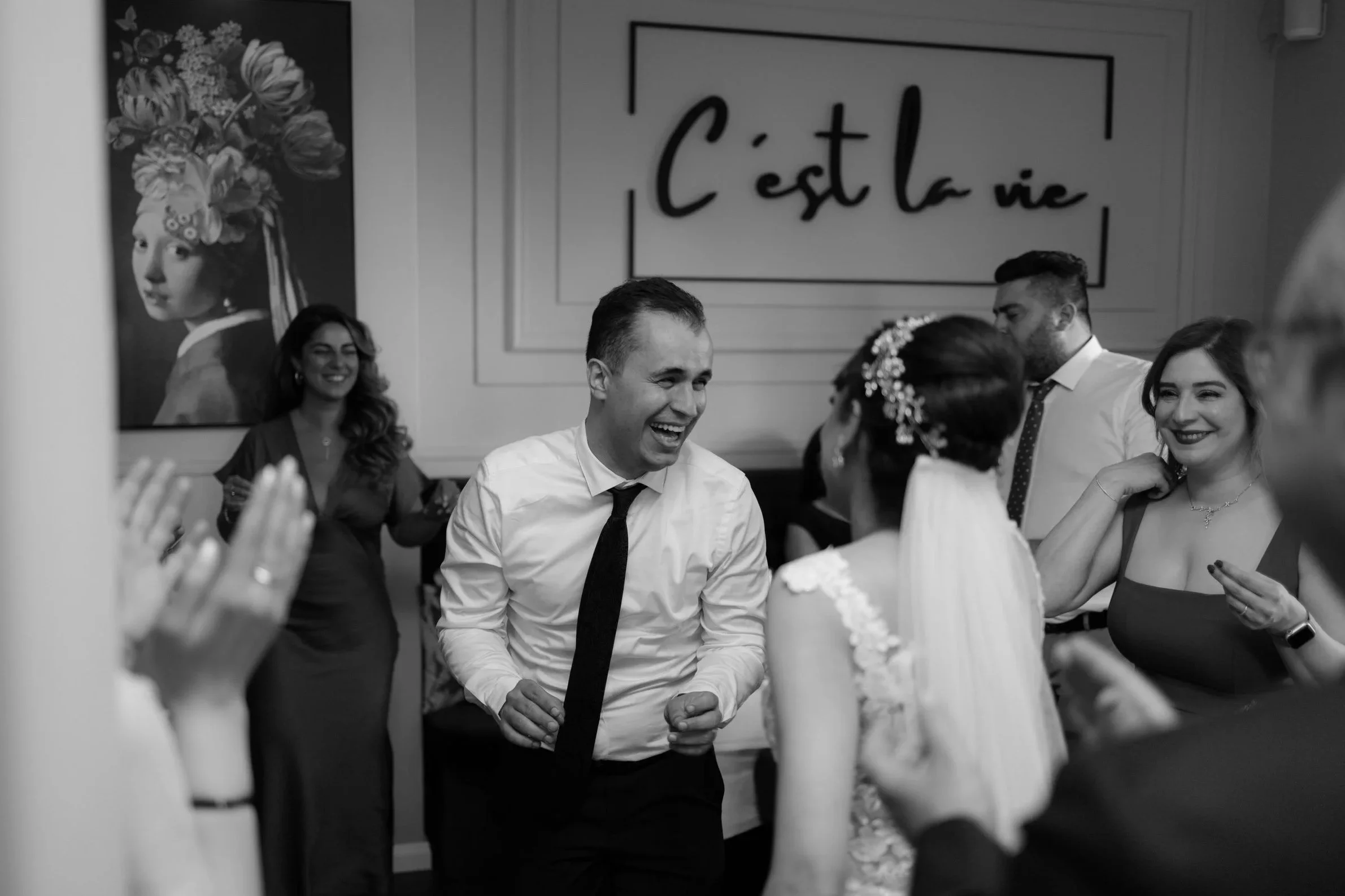 People dancing and celebrating at a wedding reception, with a man in a white shirt and black tie in the center smiling, and women around him clapping and smiling.