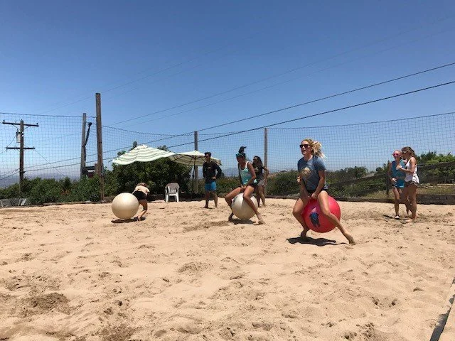 People playing with large balls on a sandy outdoor court with a fence and clear blue sky in the background.