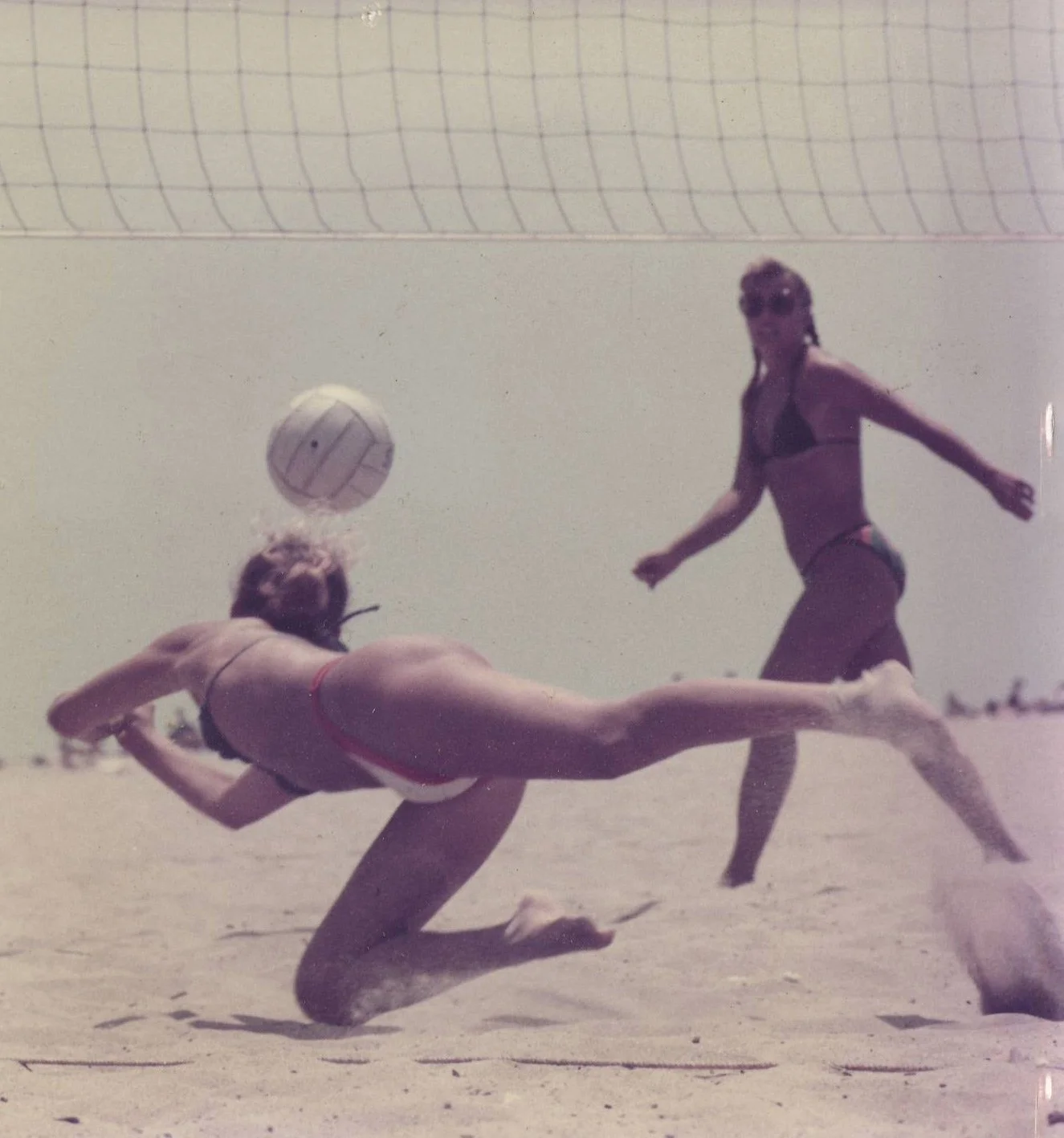 A woman diving in the sand while a woman in a bikini watches a volleyball during a beach volleyball game.