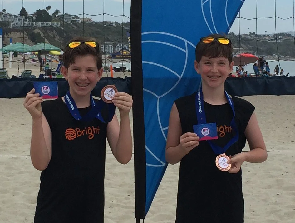 Two young boys wearing black sleeveless shirts with 'Bright' written on them, standing on a beach, each holding a medal and a card, smiling, with a volleyball court and beach umbrellas in the background.