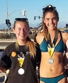 Two young women standing outdoors with medals around their necks, wearing athletic clothing and smiling at the camera.