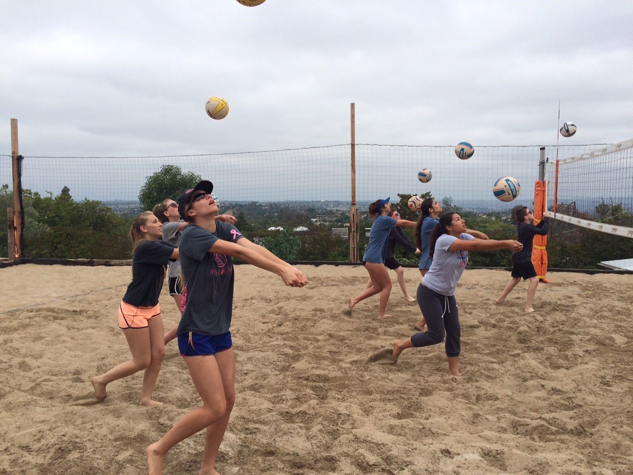 People playing beach volleyball on sand court under cloudy sky, some hitting balls near the net.